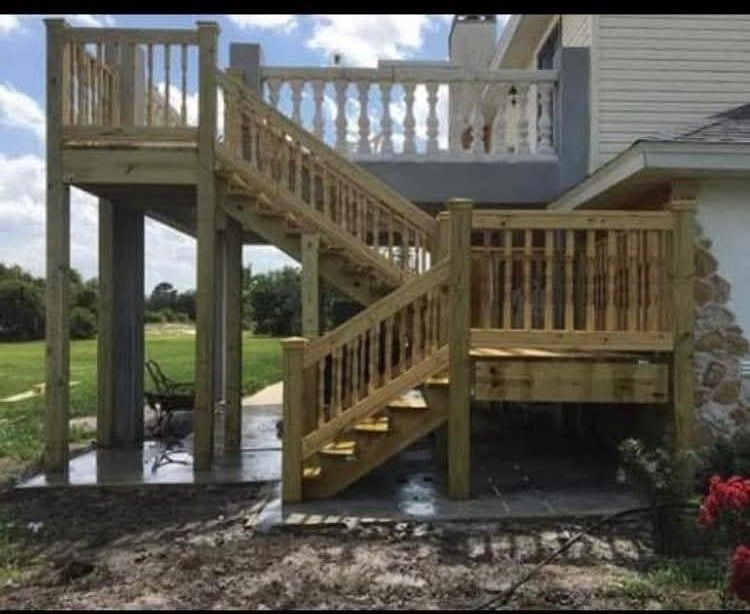 Wooden deck and stairs attached to a light-colored house, supported by posts, with a yard in the background.