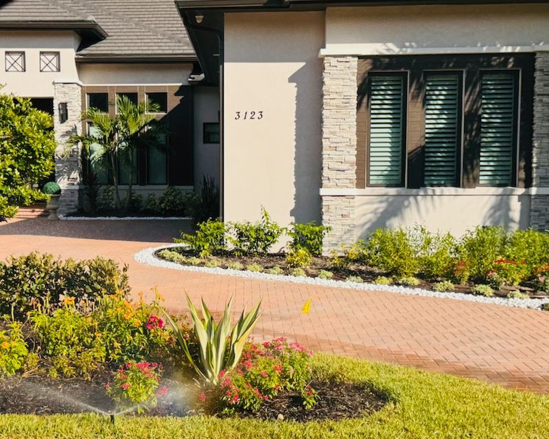 House with brick driveway, landscaping, and sprinkler system in use.