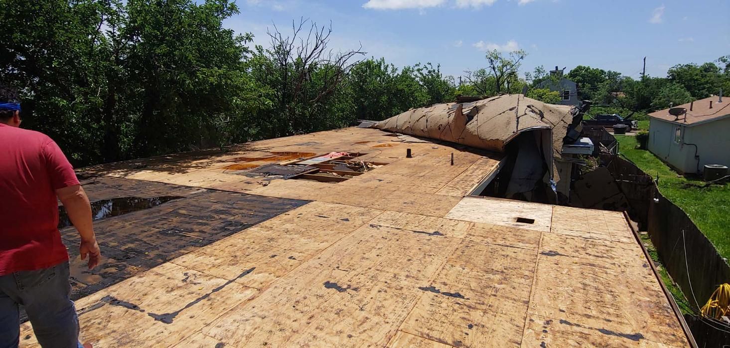 Rooftop with damaged wooden panels and a collapsed section, trees and houses in the background