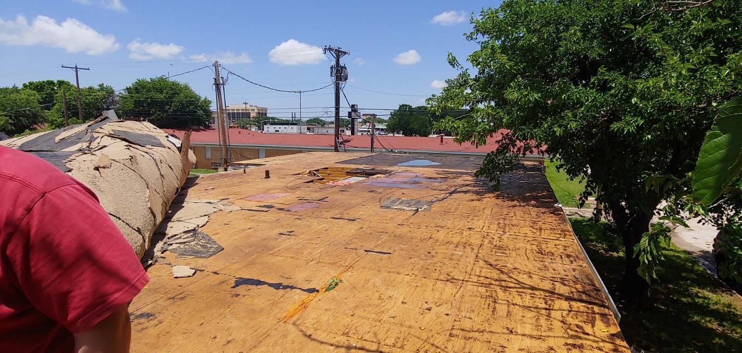 Damaged yellow-orange road surface with cracks and patches beside a tree-lined street under a blue sky