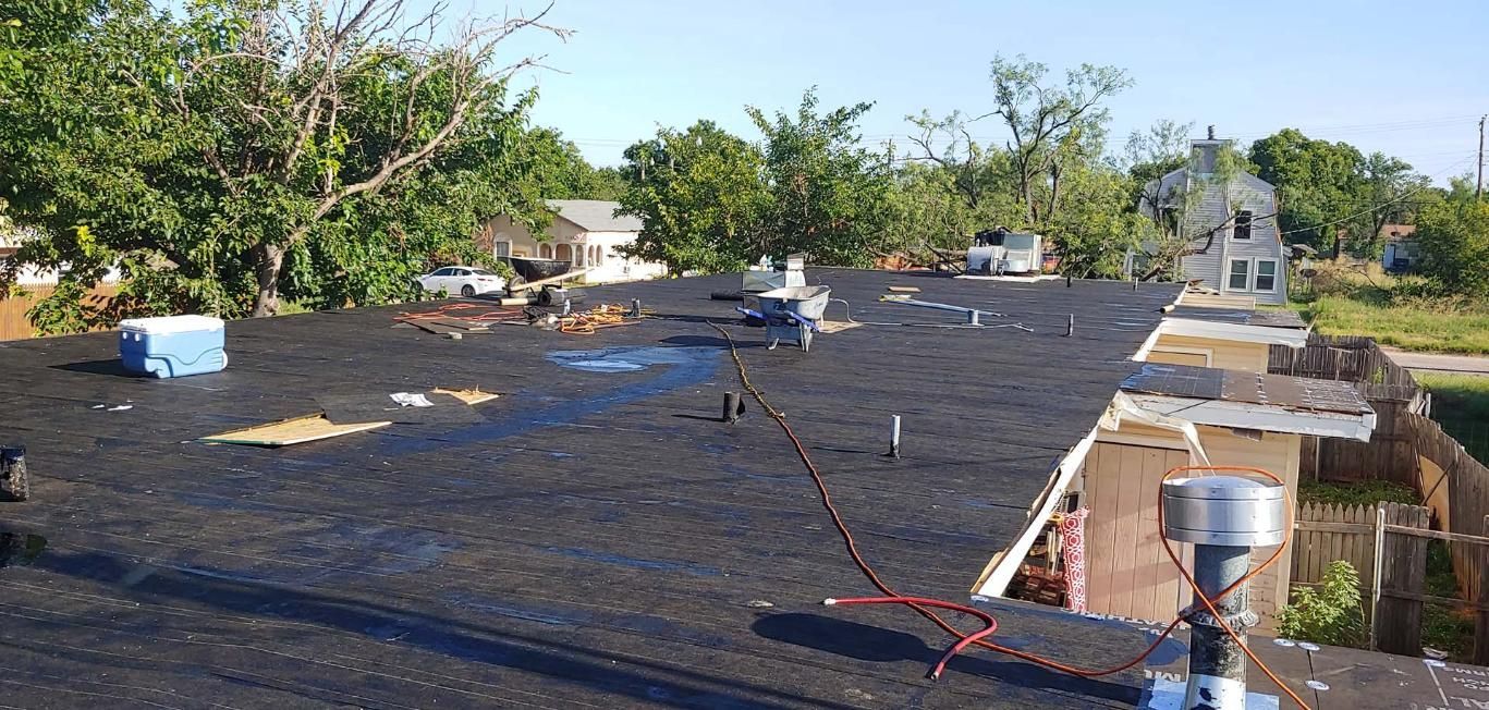 Flat rooftop covered in black waterproofing, with vents and tools near trees and blue sky