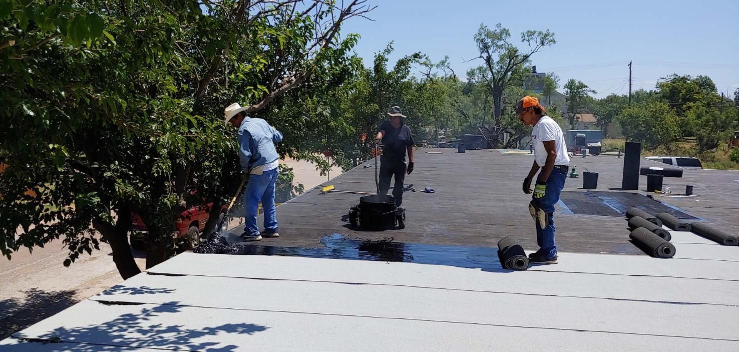 Workers installing black roofing material on a flat roof under a bright sky