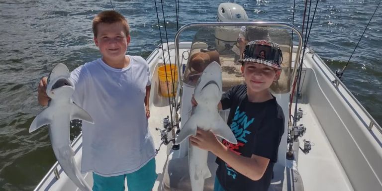 Two young boys are standing on a boat holding sharks.