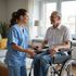Woman in blue scrubs assists a man in a wheelchair; they are smiling and talking indoors.