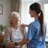 Woman with gray hair smiles while a person in blue scrubs puts a hand on her shoulder.