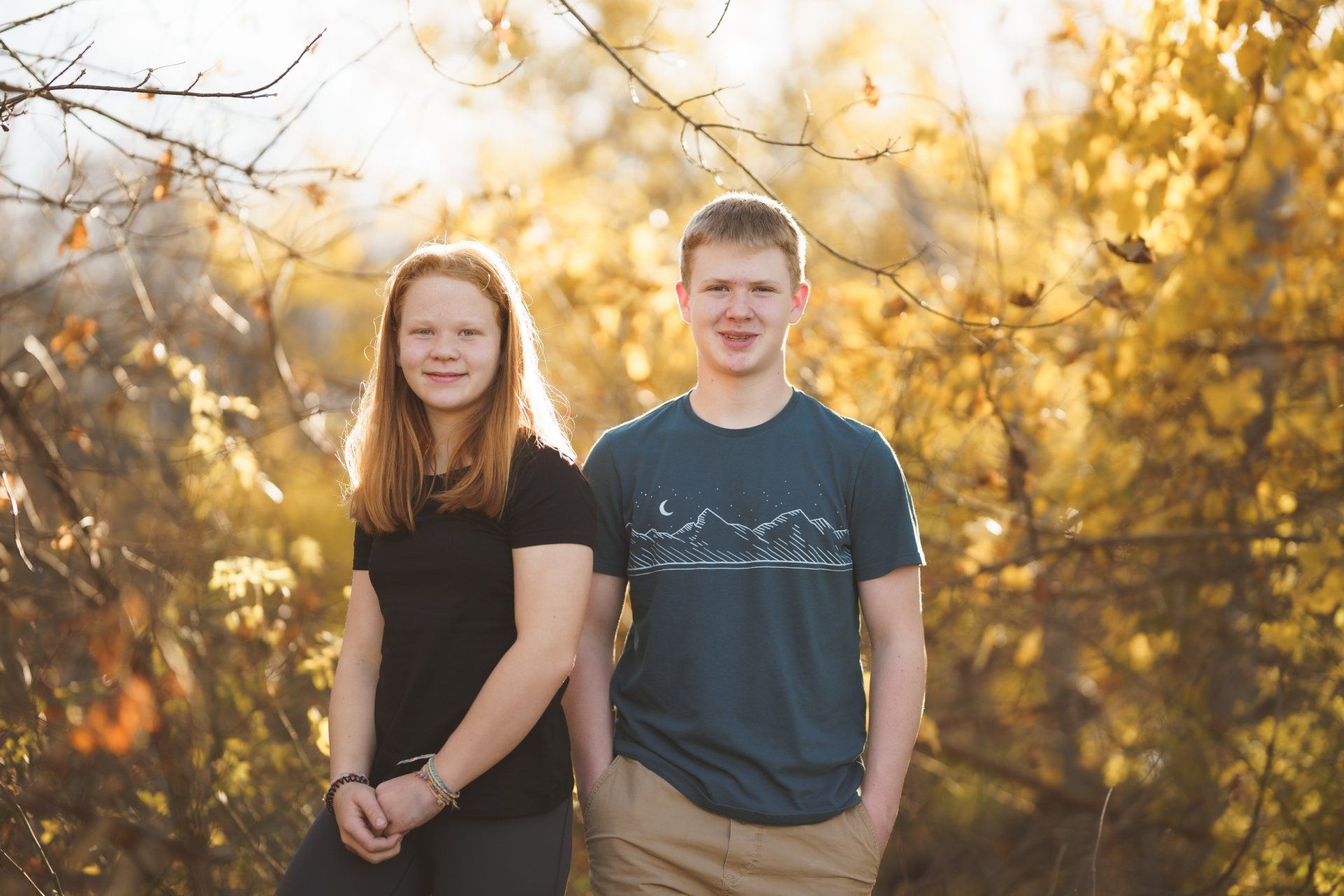 A boy and a girl are standing next to each other in front of trees.