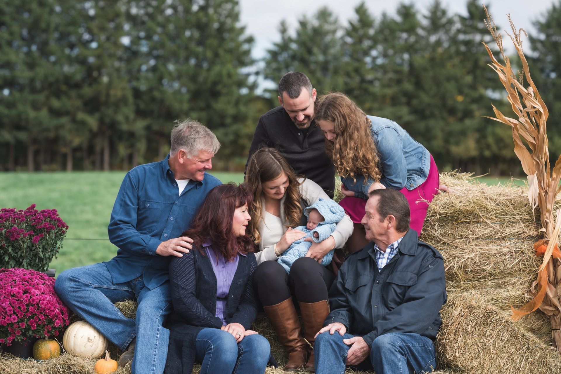 A family is sitting on a bale of hay looking at a baby.