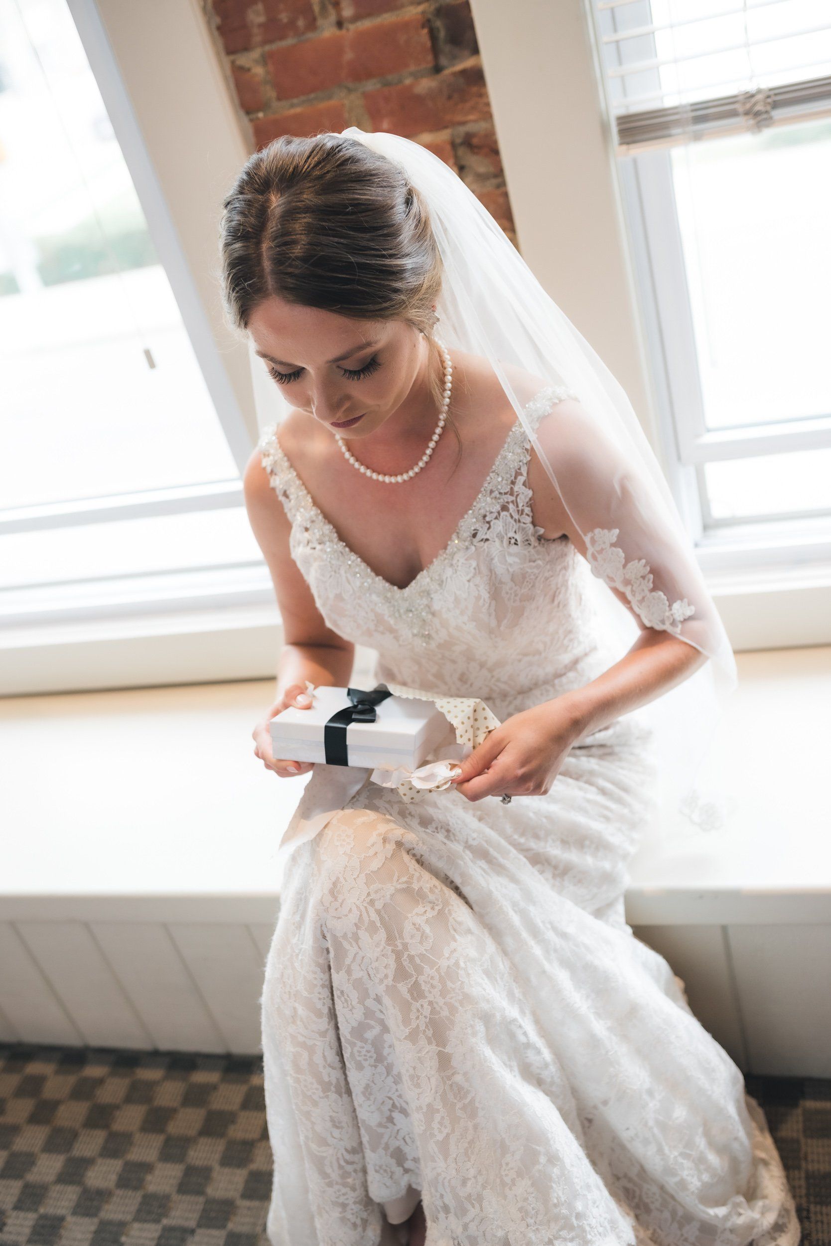 A bride in a wedding dress is sitting on a window sill holding a gift box.