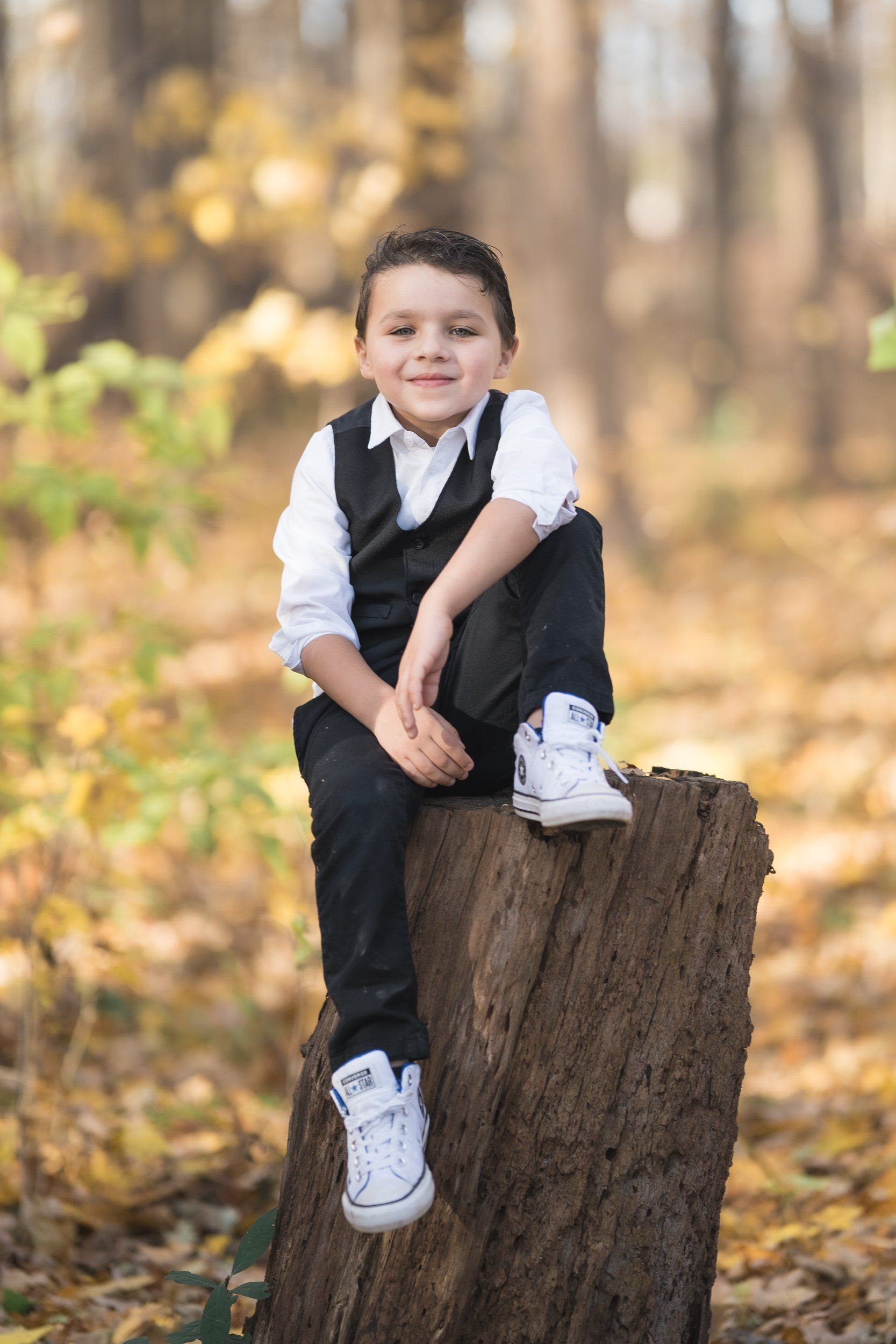 A young boy is sitting on a tree stump in the woods.
