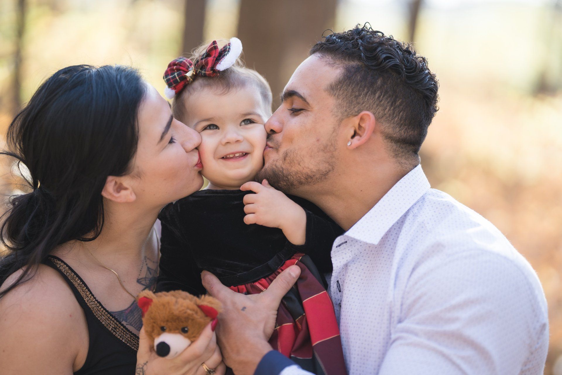 A man and woman are kissing a little girl on the cheek while holding a teddy bear.