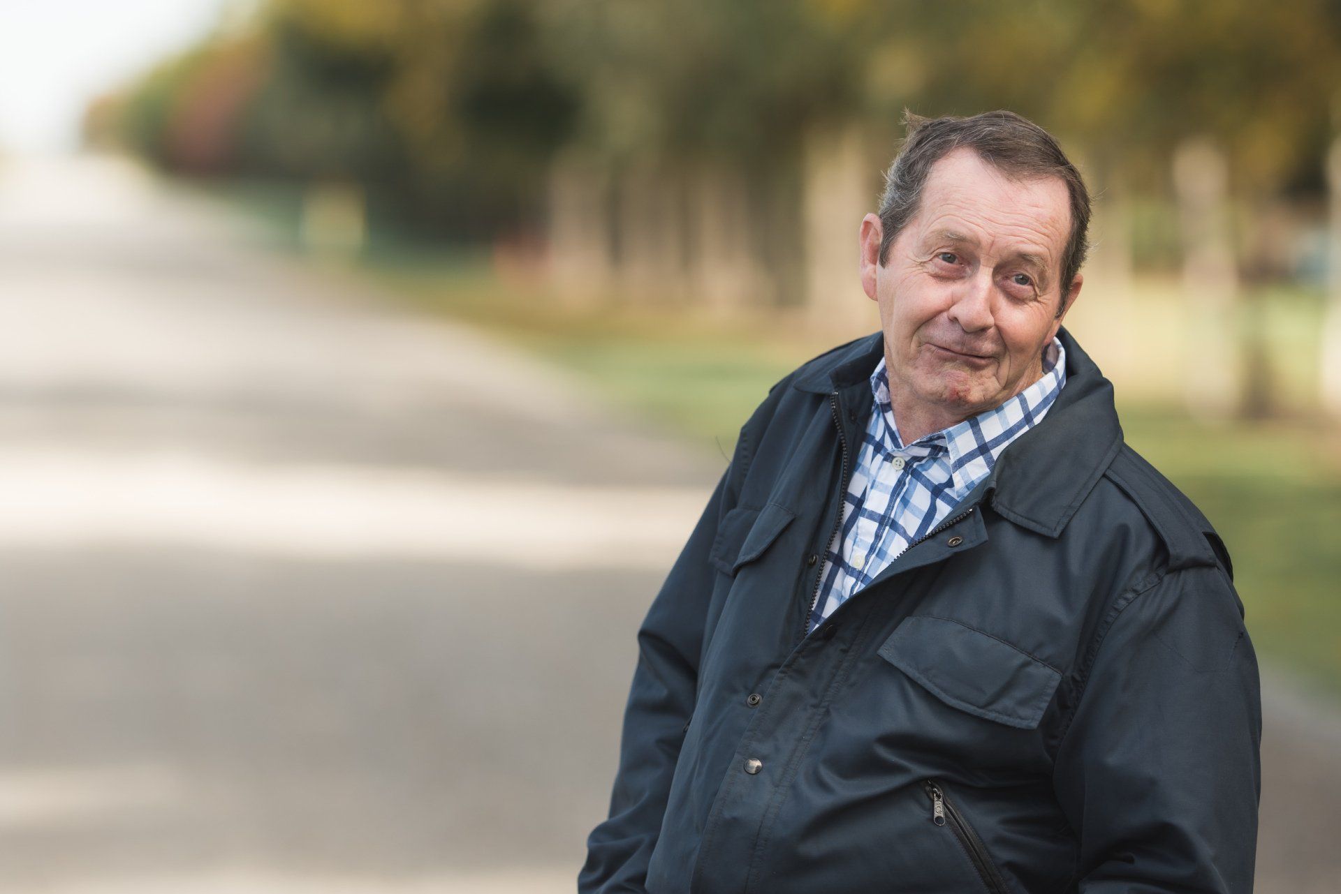 A man in a black jacket and plaid shirt is standing on the side of a road.