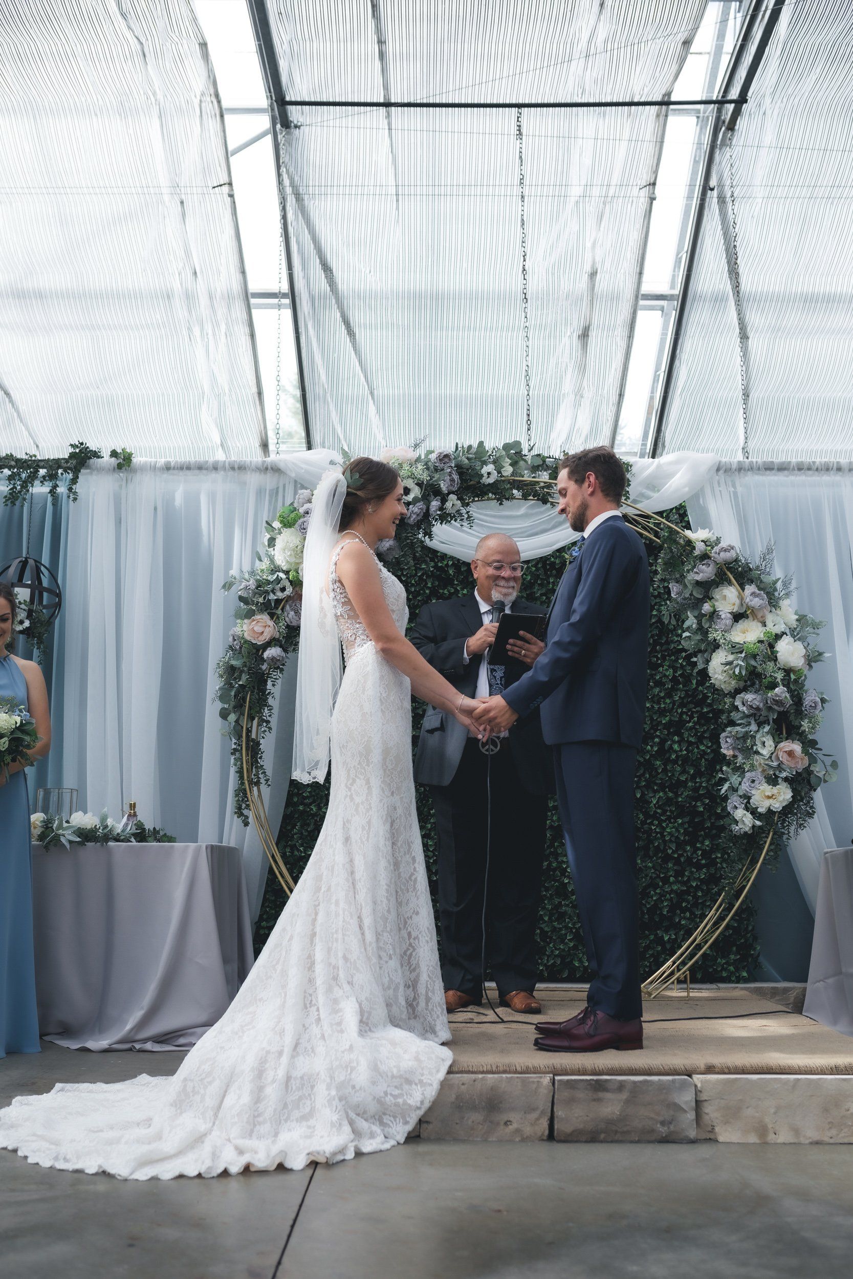 A bride and groom are holding hands during their wedding ceremony in a greenhouse.