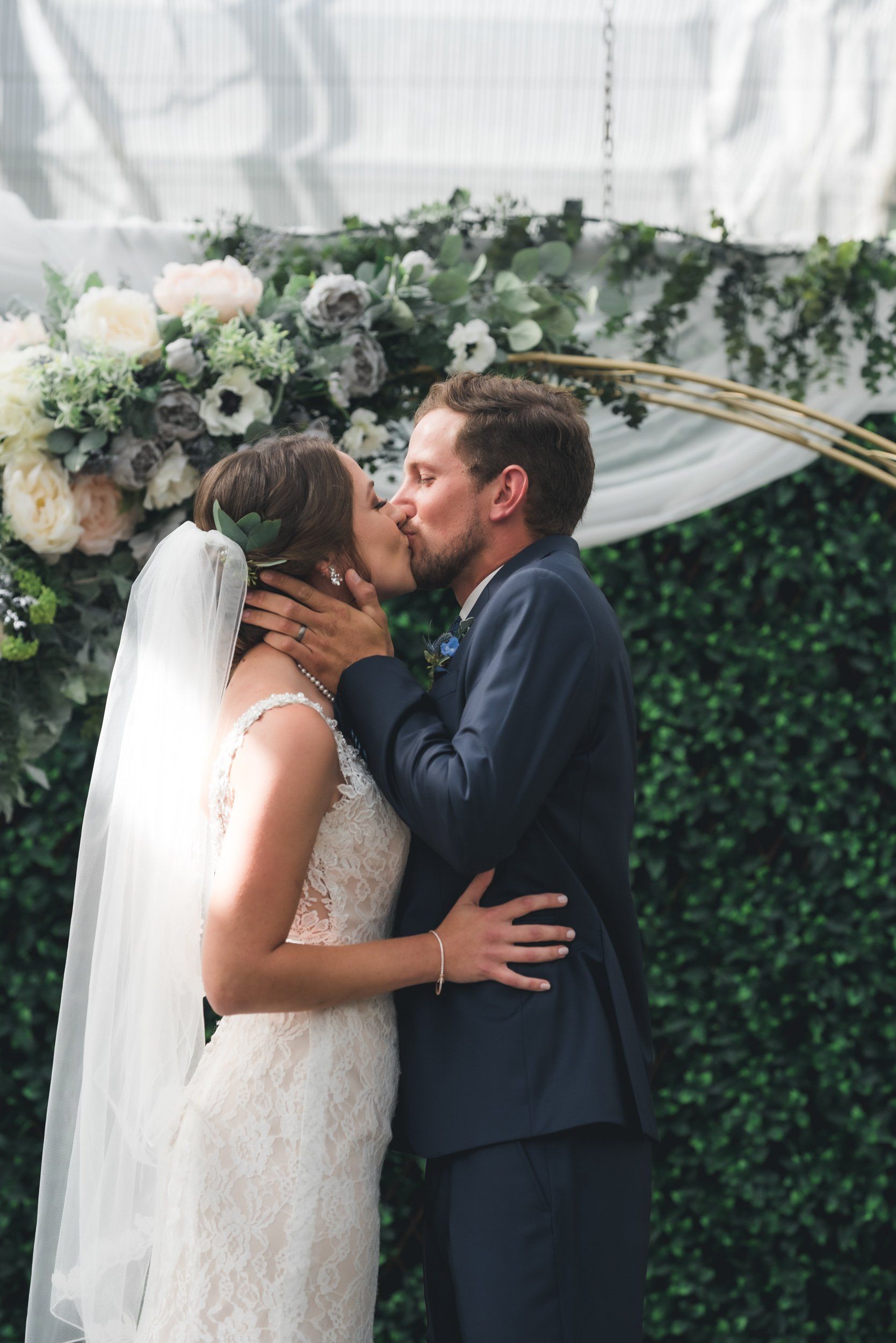 A bride and groom are kissing under a floral arch at their wedding ceremony.