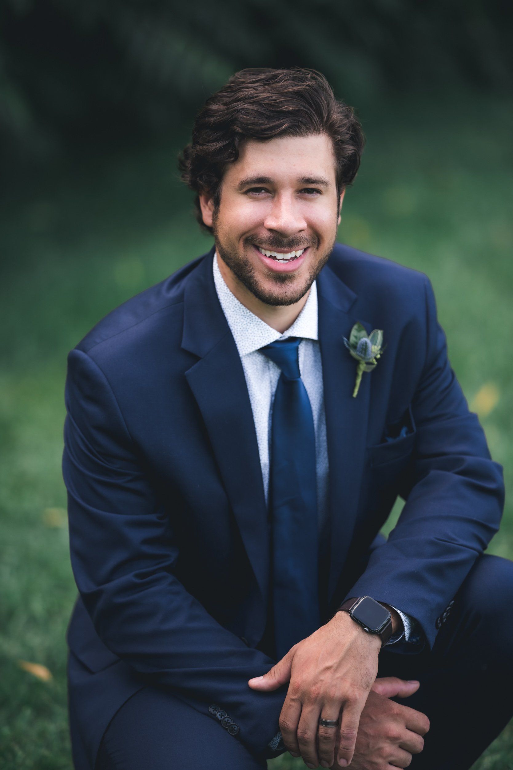 A man in a suit and tie is kneeling down in the grass and smiling.