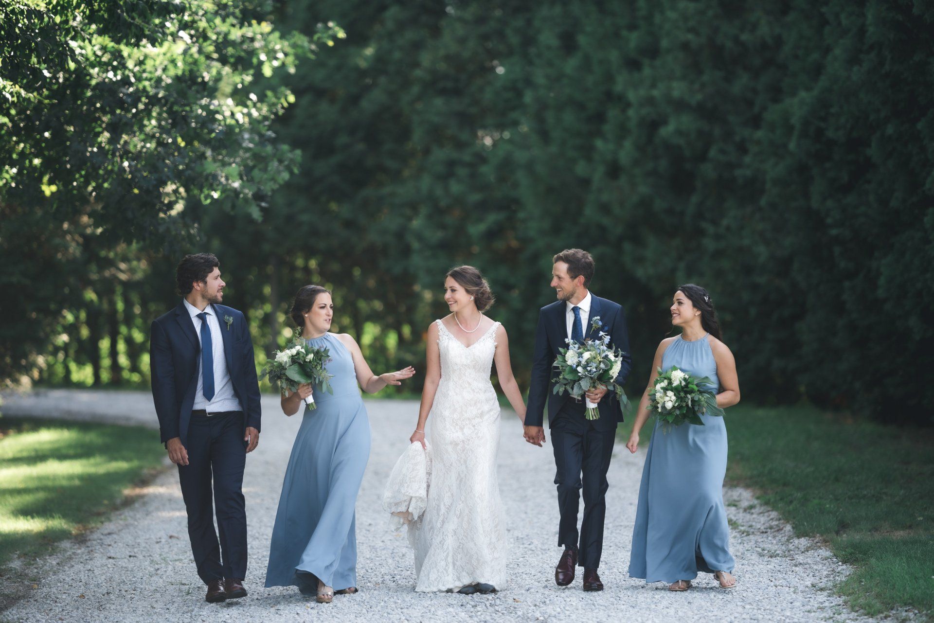 A bride and groom are walking down a gravel road with their bridesmaids.