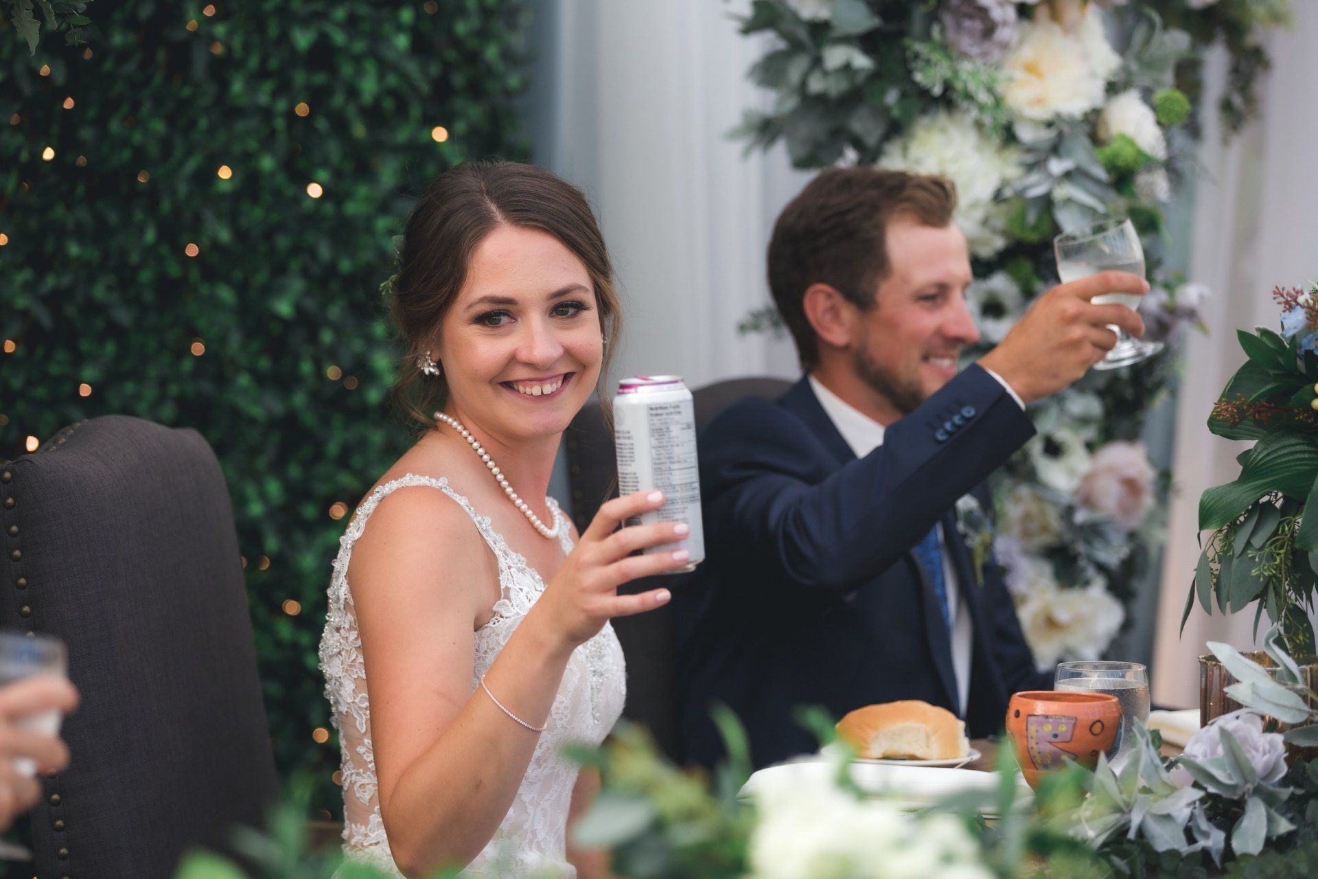 A bride and groom are toasting at their wedding reception while the bride is holding a can of beer.