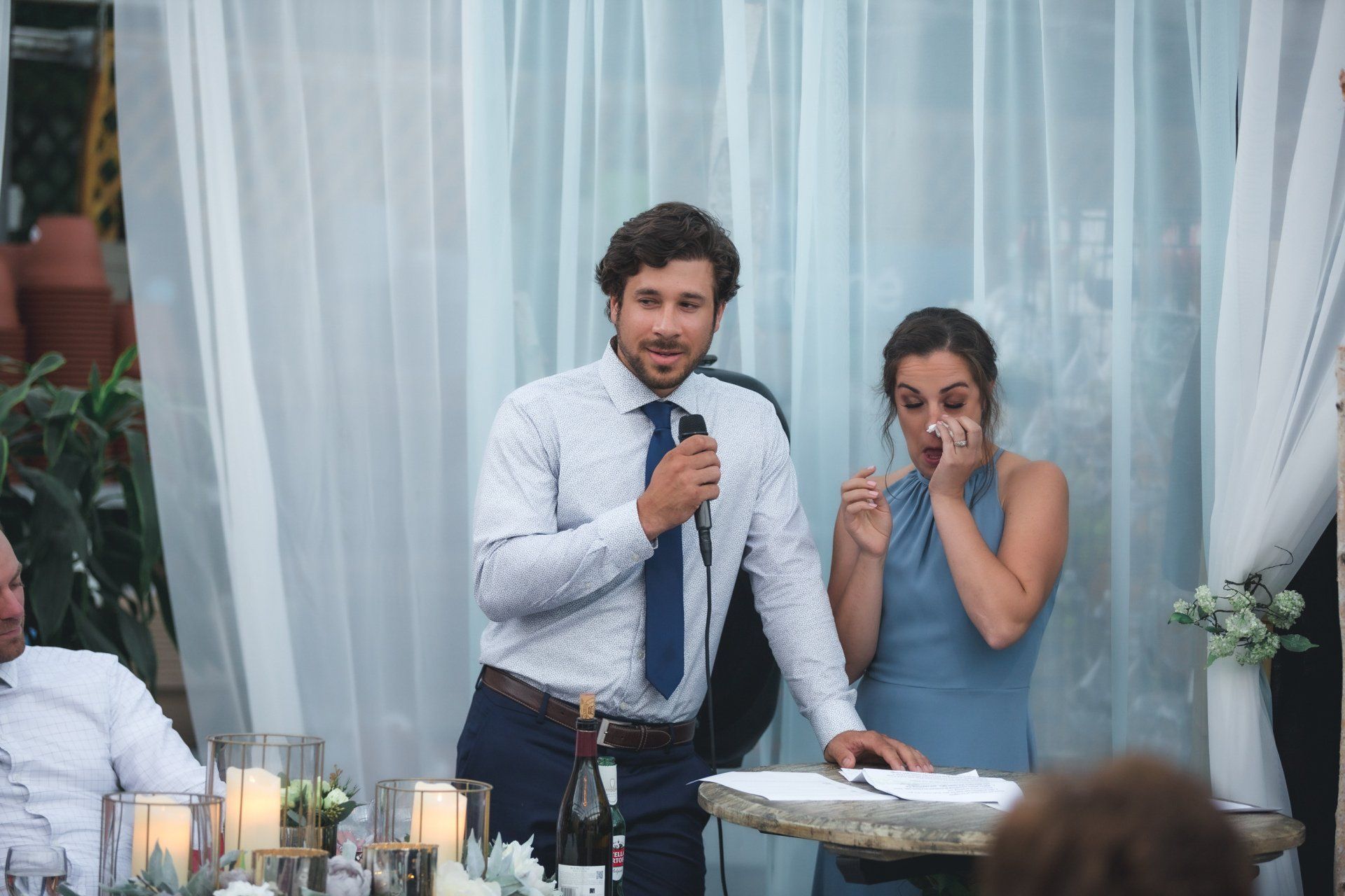 A man is giving a speech at a wedding reception while a woman wipes her eyes.