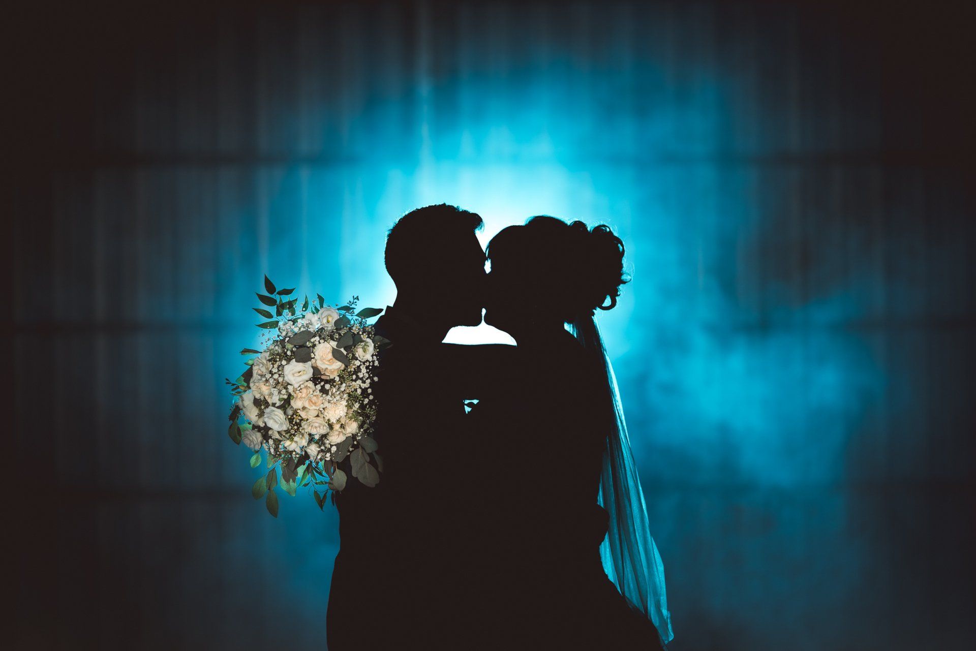 A bride and groom are kissing in front of a blue background . the bride is holding a bouquet of flowers.