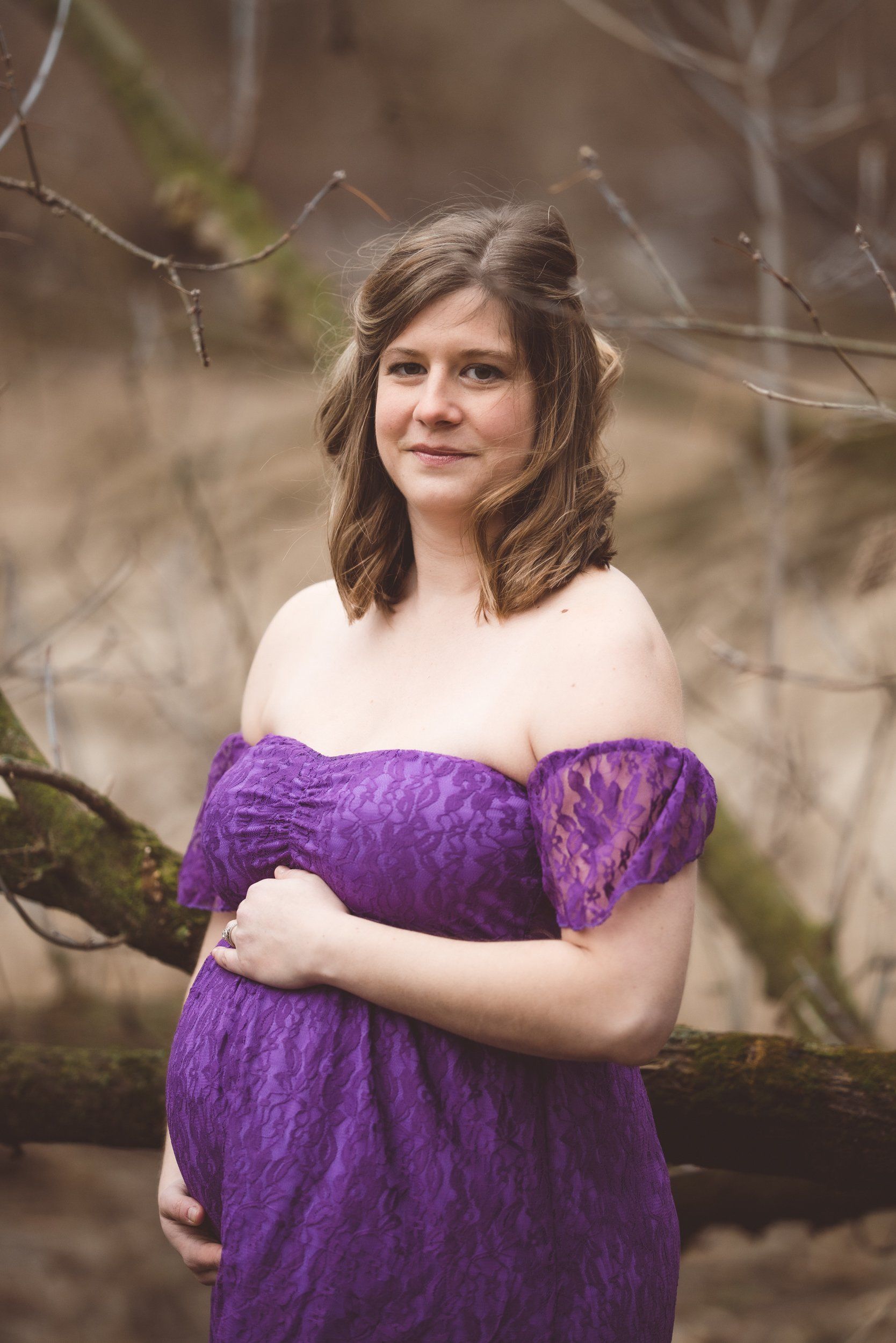 A pregnant woman in a purple dress is standing next to a tree holding her belly.