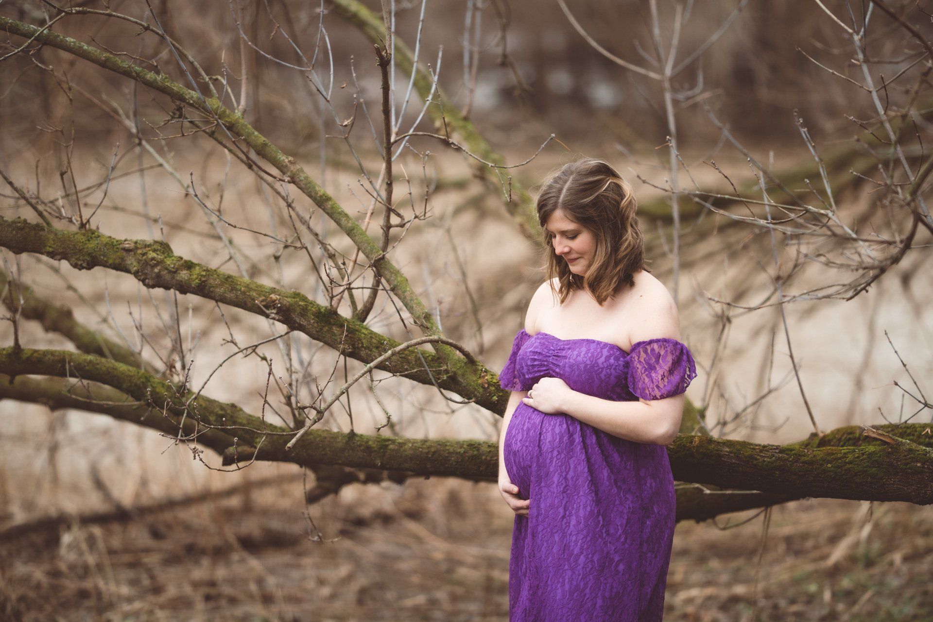 A pregnant woman in a purple dress is standing next to a tree.