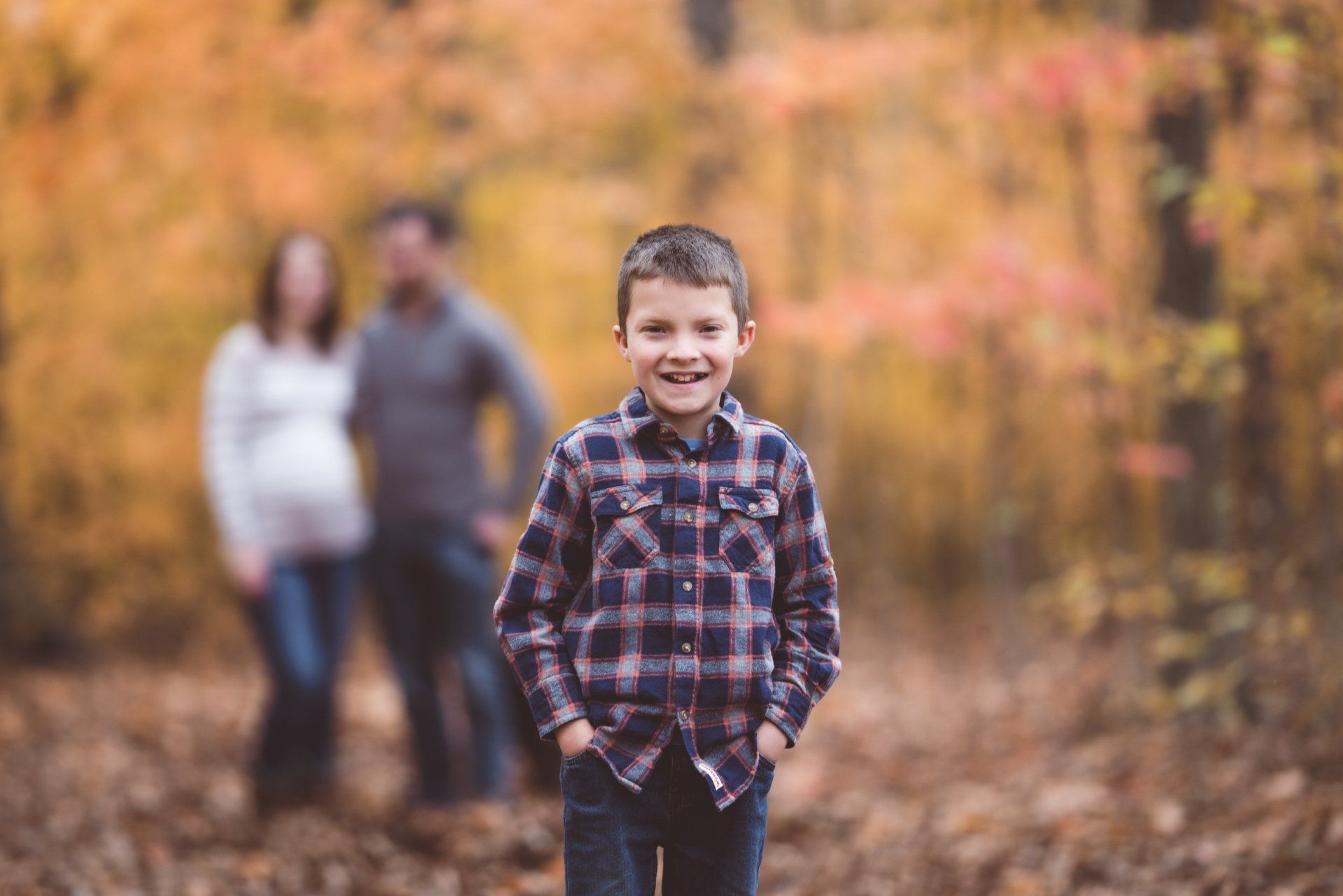A young boy is standing in the woods with his parents in the background.