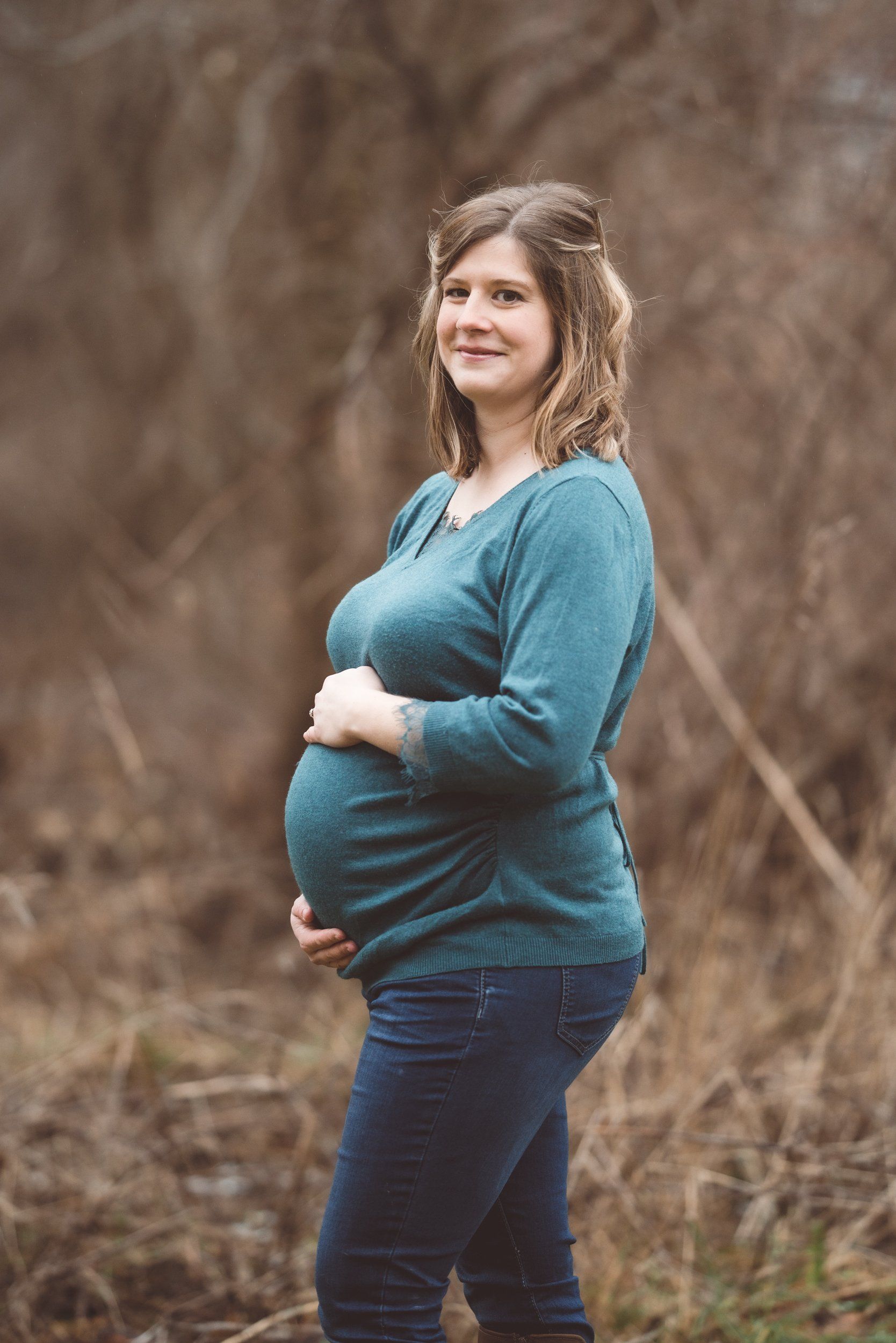 A pregnant woman is standing in a field holding her belly.