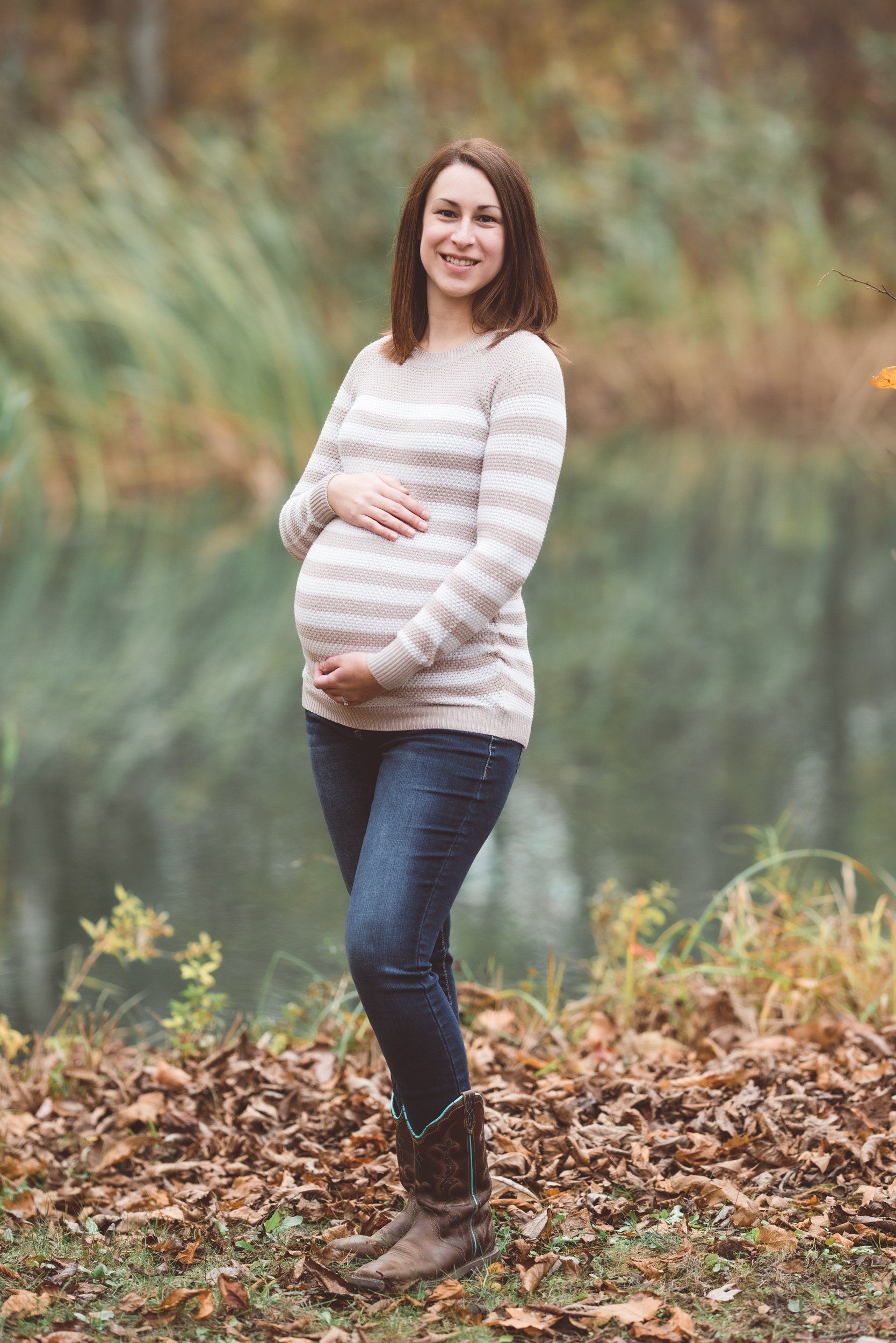 A pregnant woman is standing in front of a body of water holding her belly.