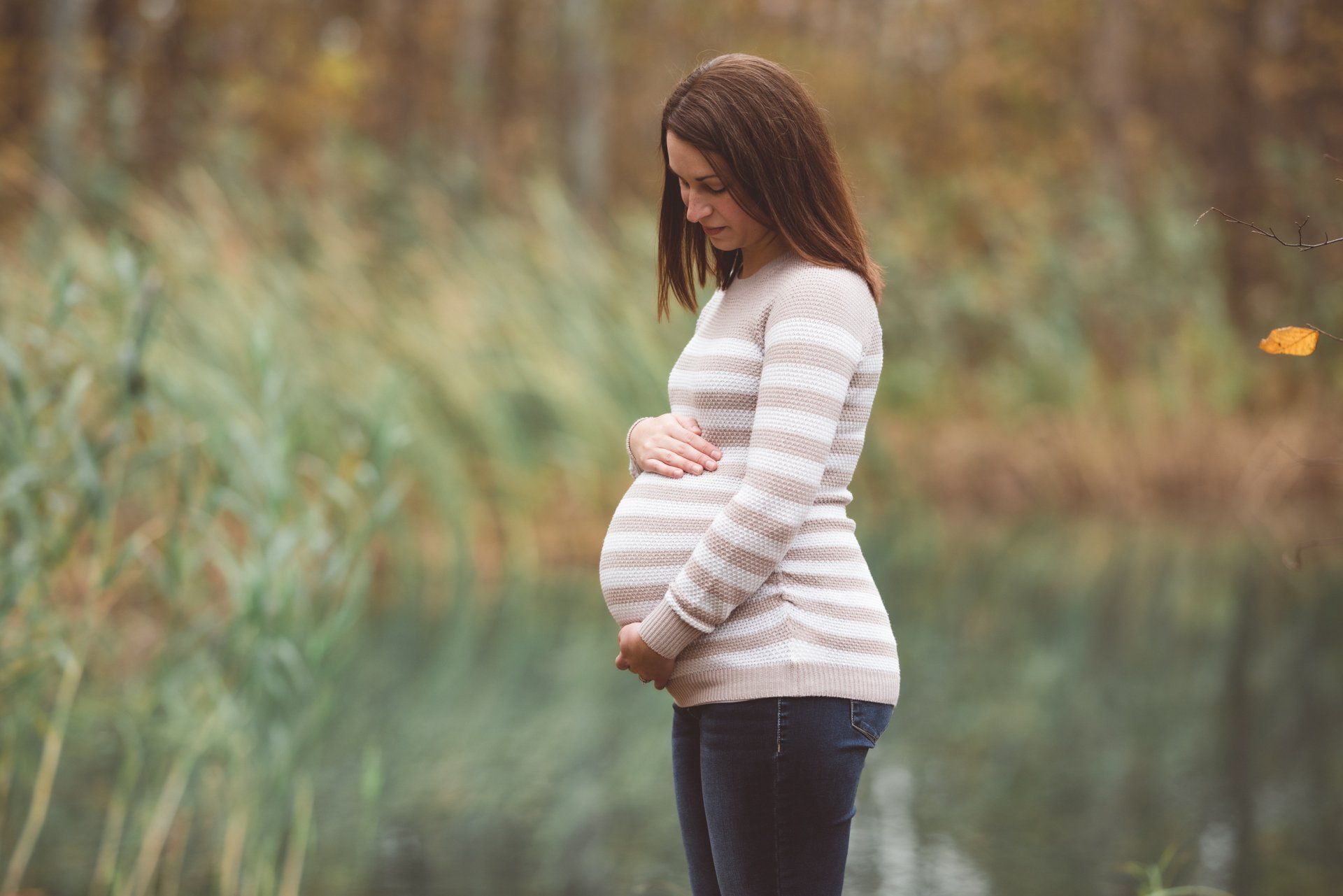 A pregnant woman is standing in front of a body of water holding her belly.