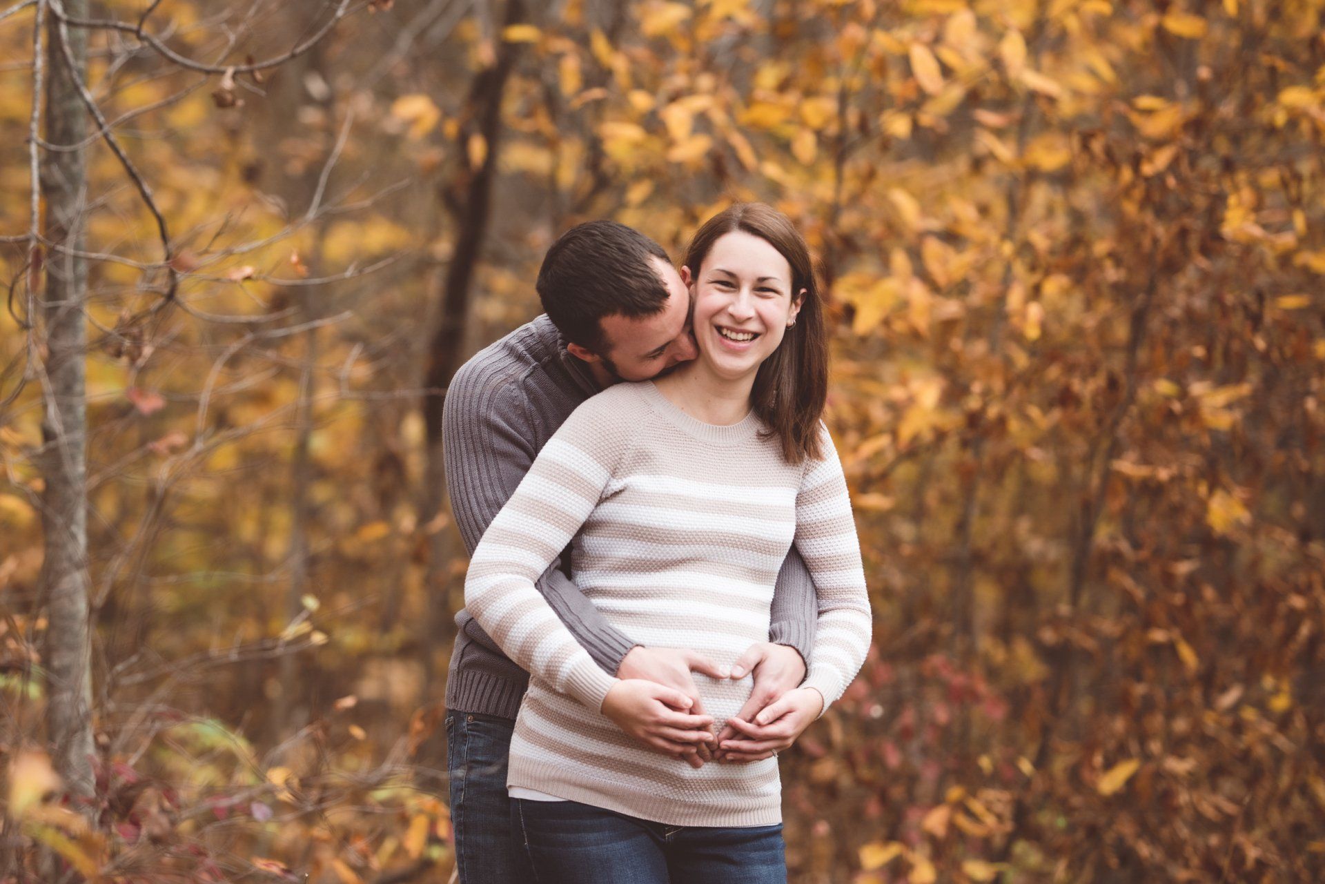 A man is kissing a pregnant woman on the cheek in the woods.