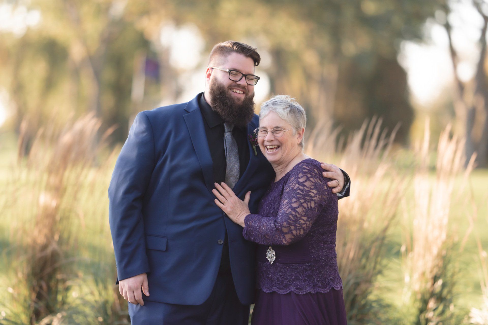 A man in a suit and a woman in a purple dress are standing next to each other in a field.