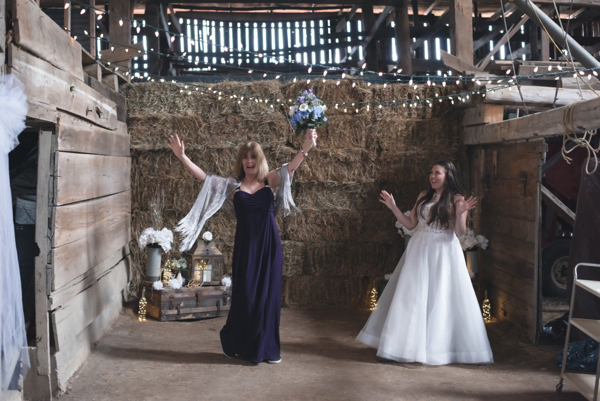 A bride and her bridesmaid are standing in a barn holding a bouquet of flowers.