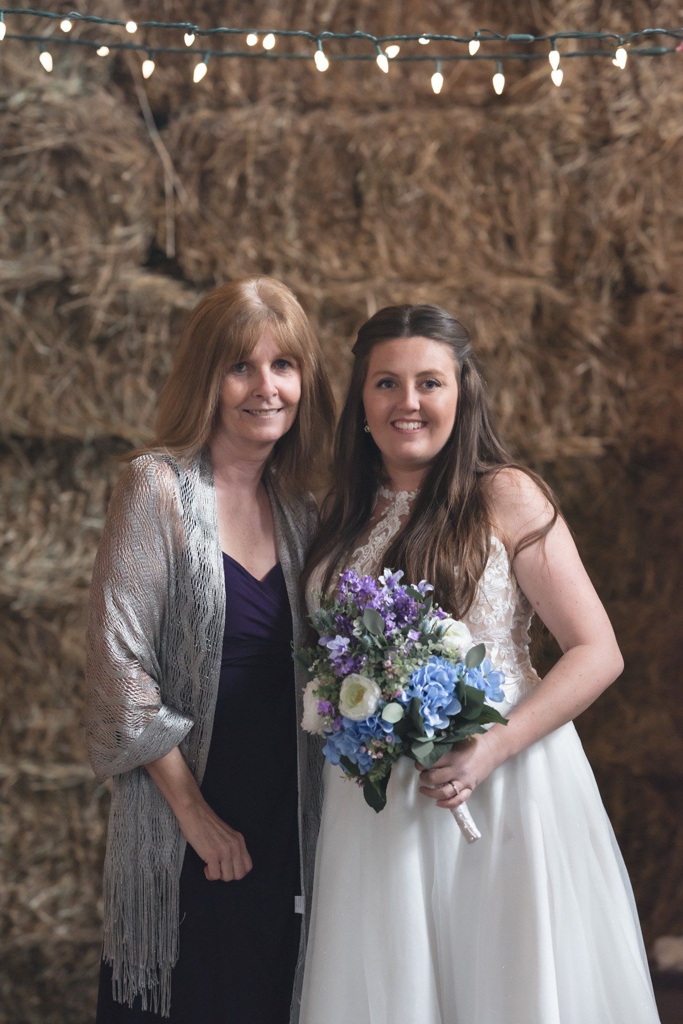 A bride and her mother are posing for a picture in front of a hay bale wall.