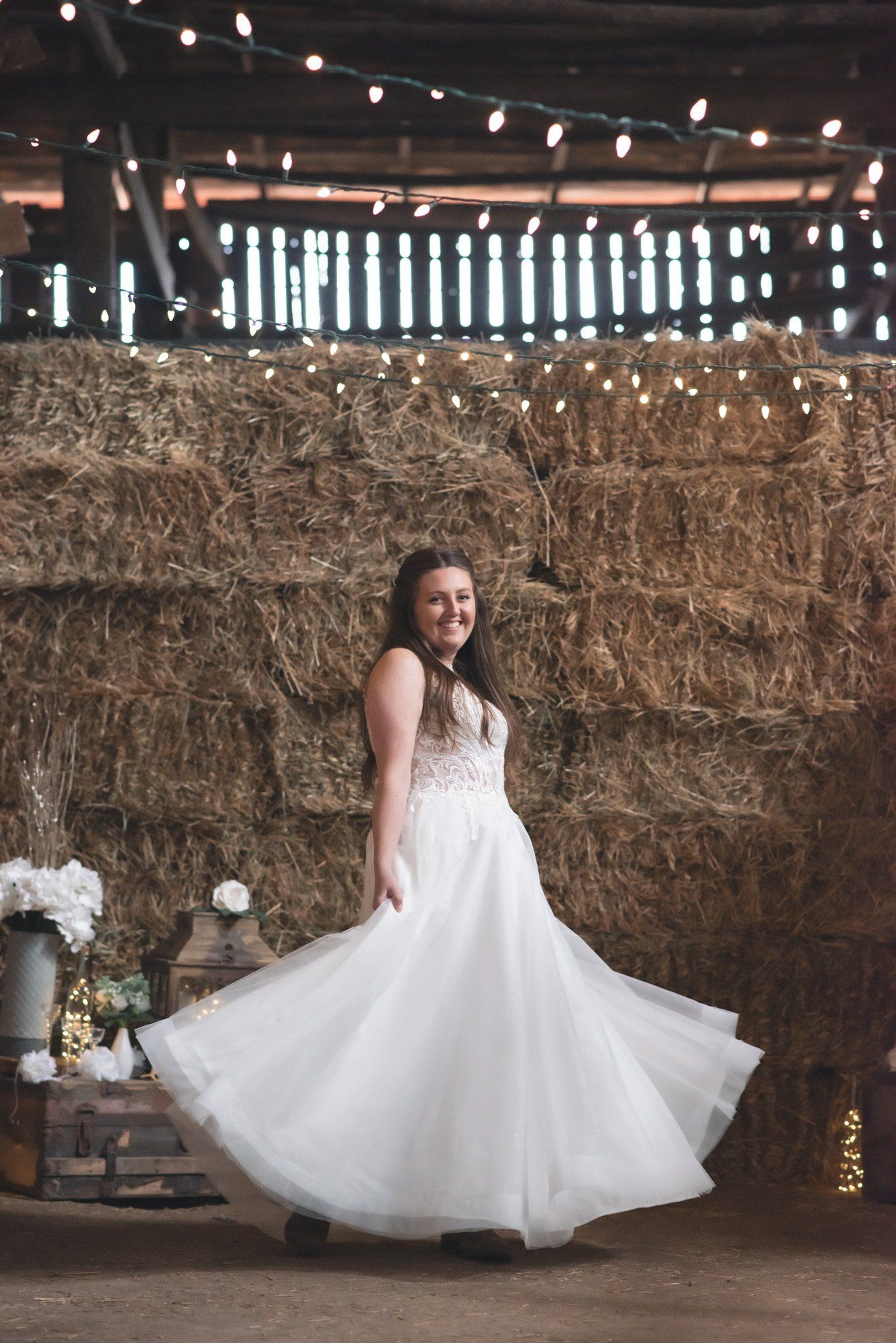 A woman in a wedding dress is dancing in front of a hay bale.