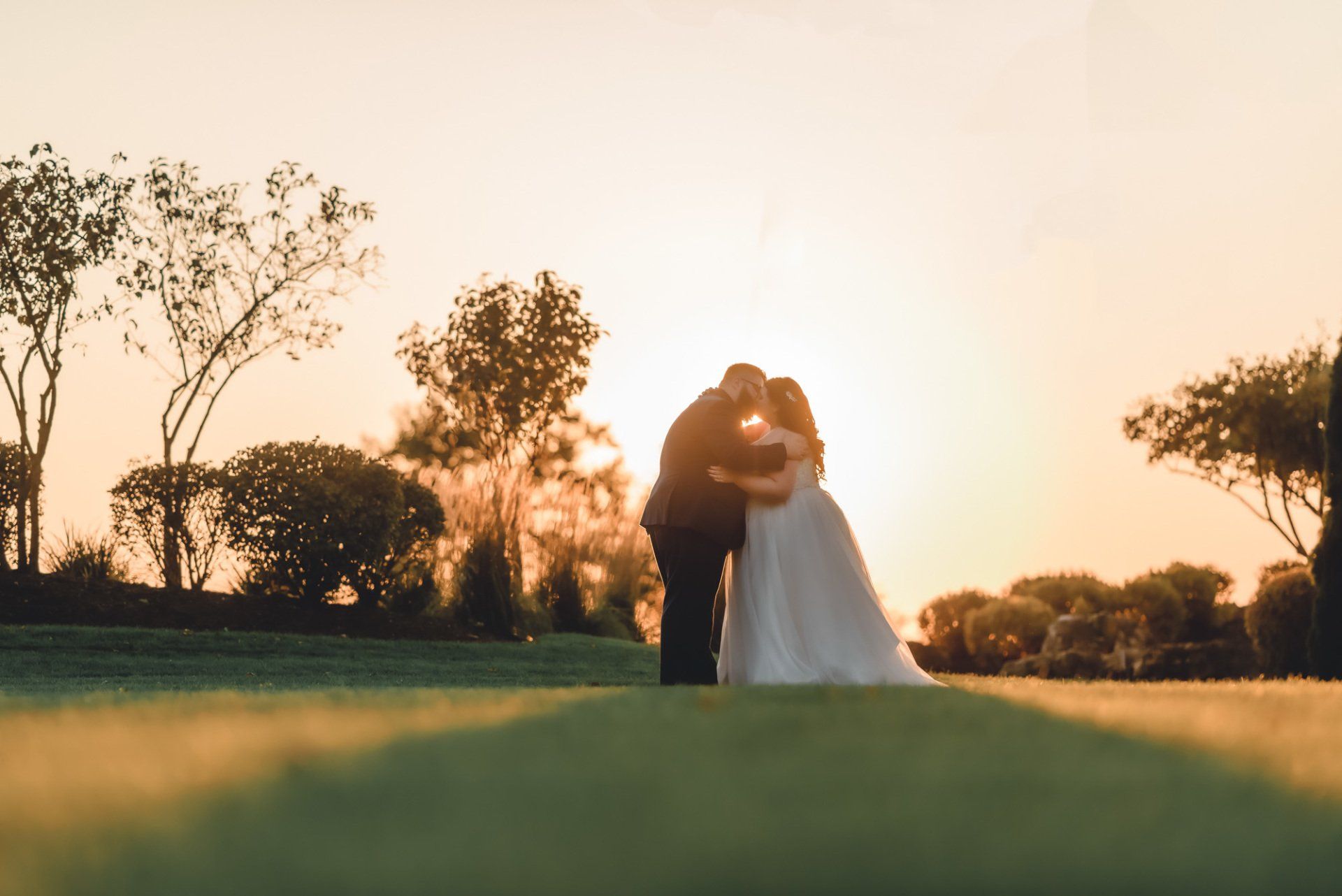 A bride and groom are kissing in a field at sunset.