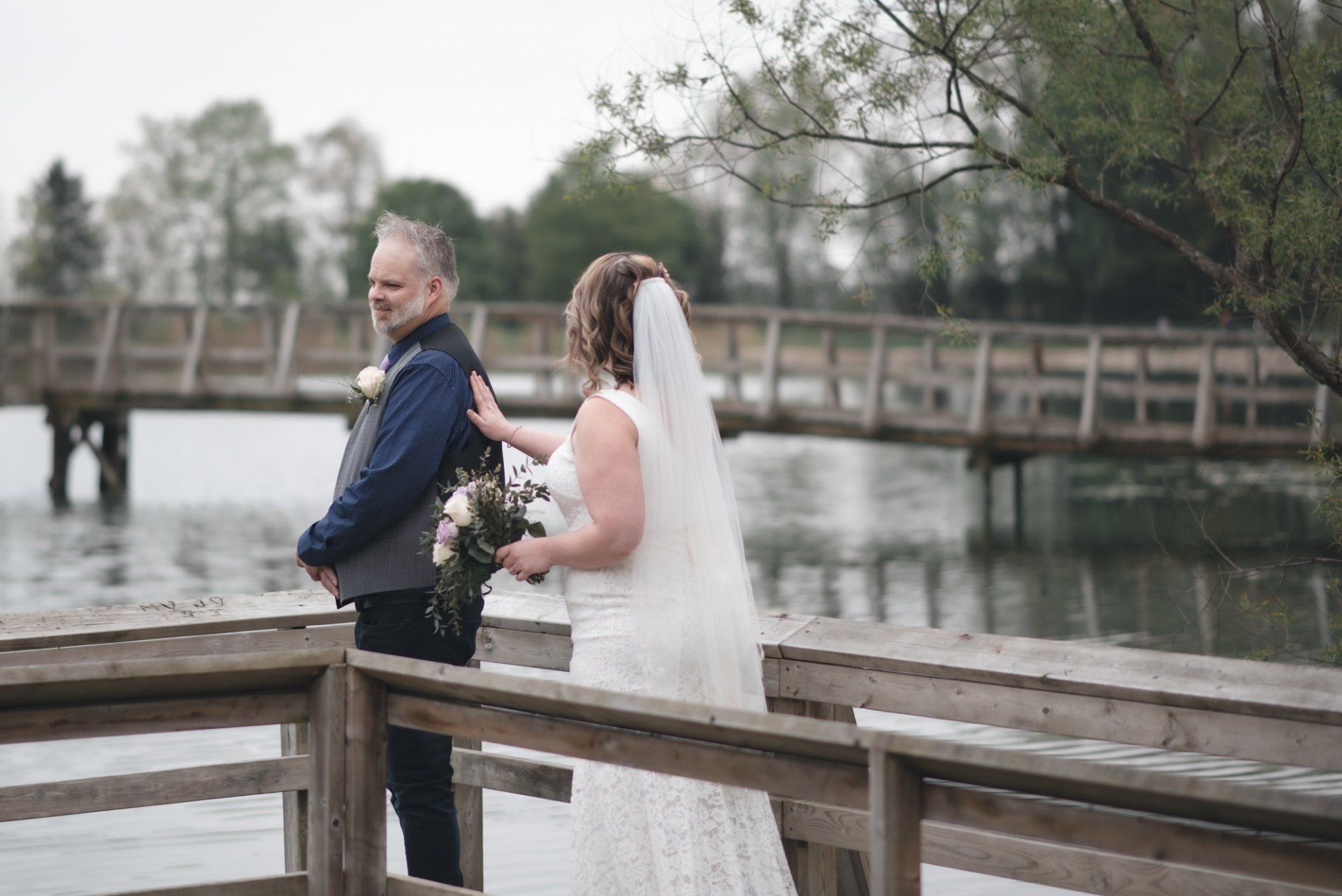 A bride and groom are standing on a dock overlooking a lake.