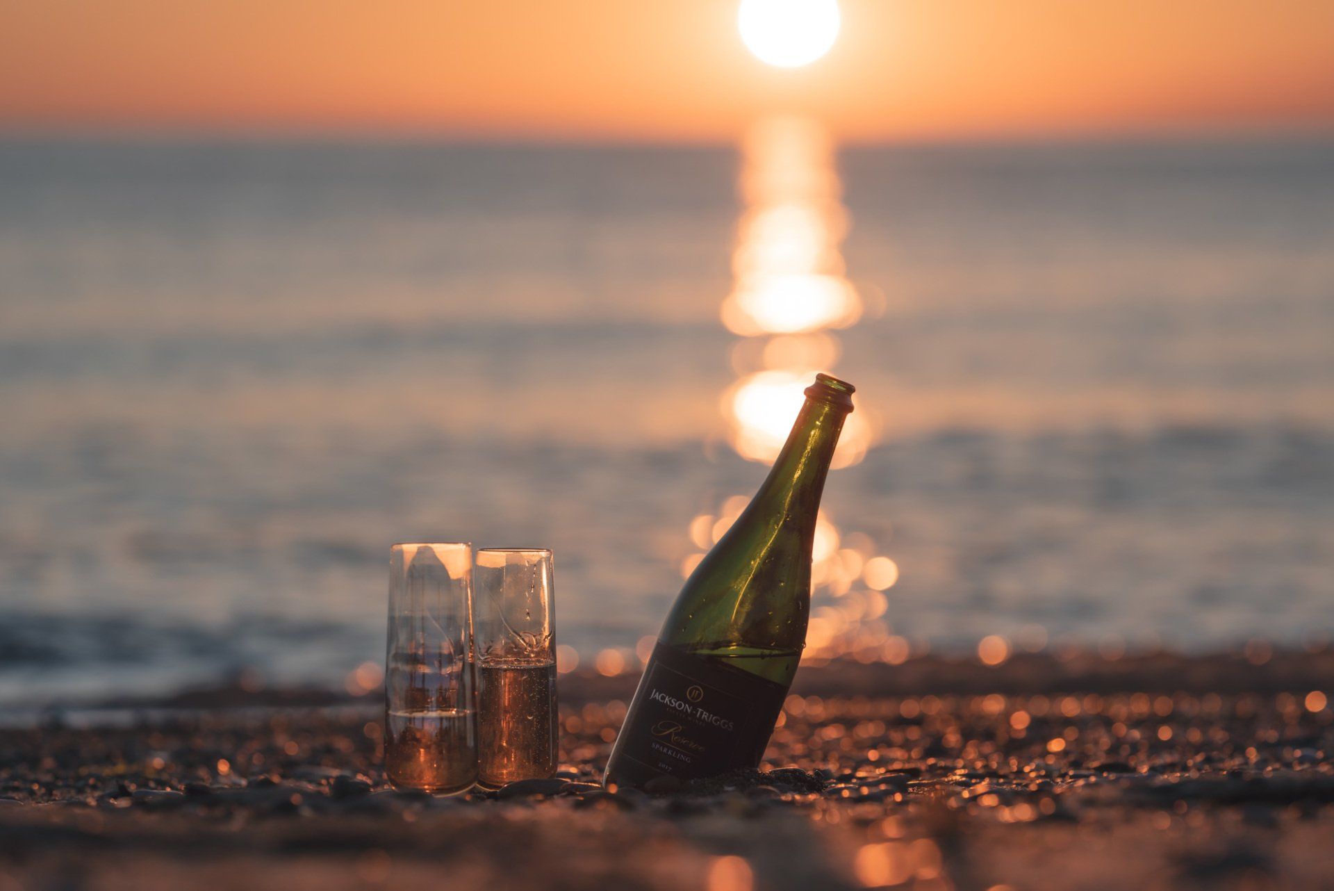 A bottle of champagne and two glasses on the beach at sunset.