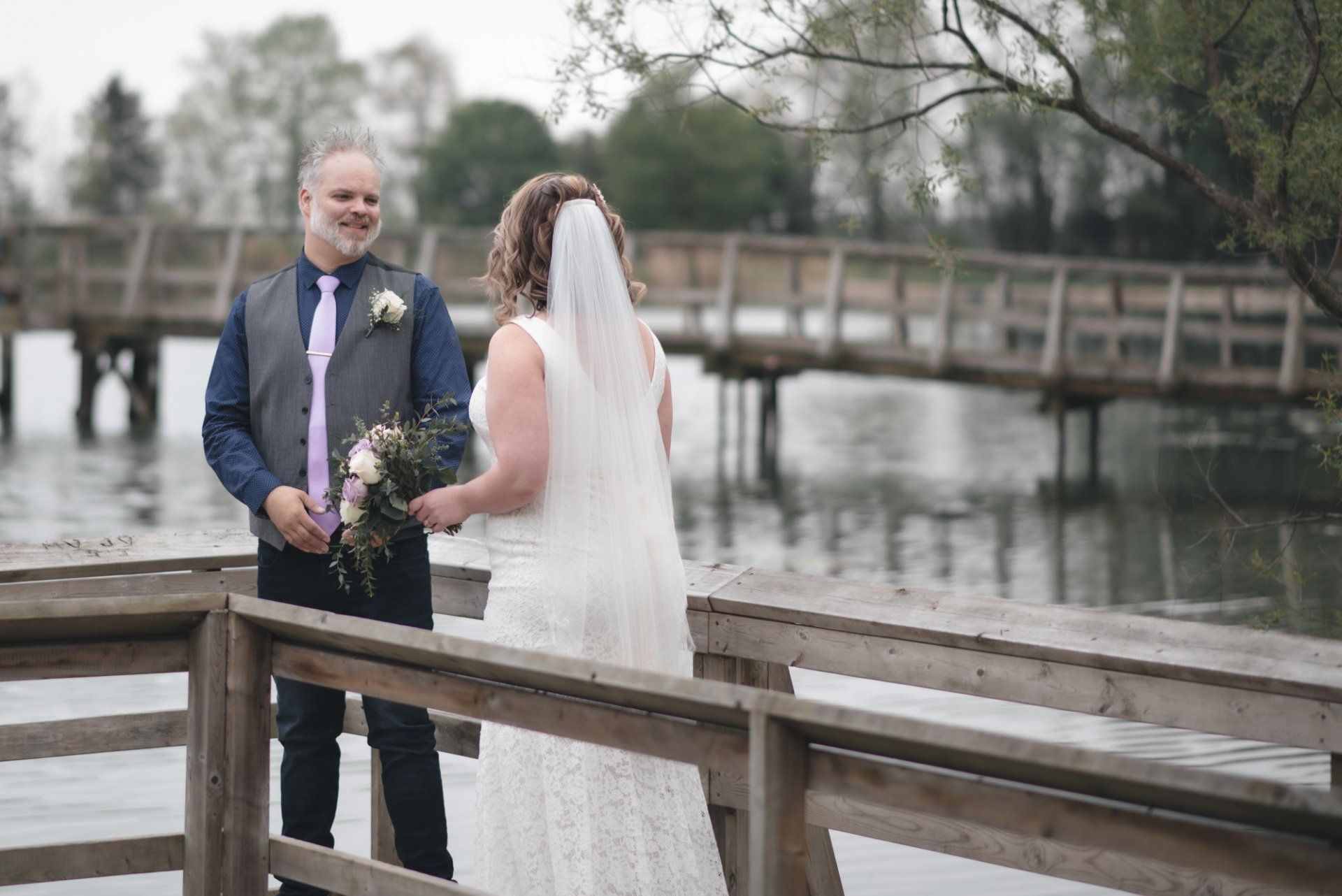 A bride and groom are standing on a wooden dock overlooking a lake.