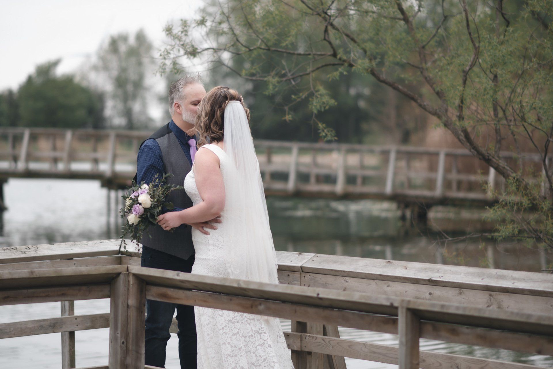 A bride and groom are kissing on a wooden bridge over a lake.