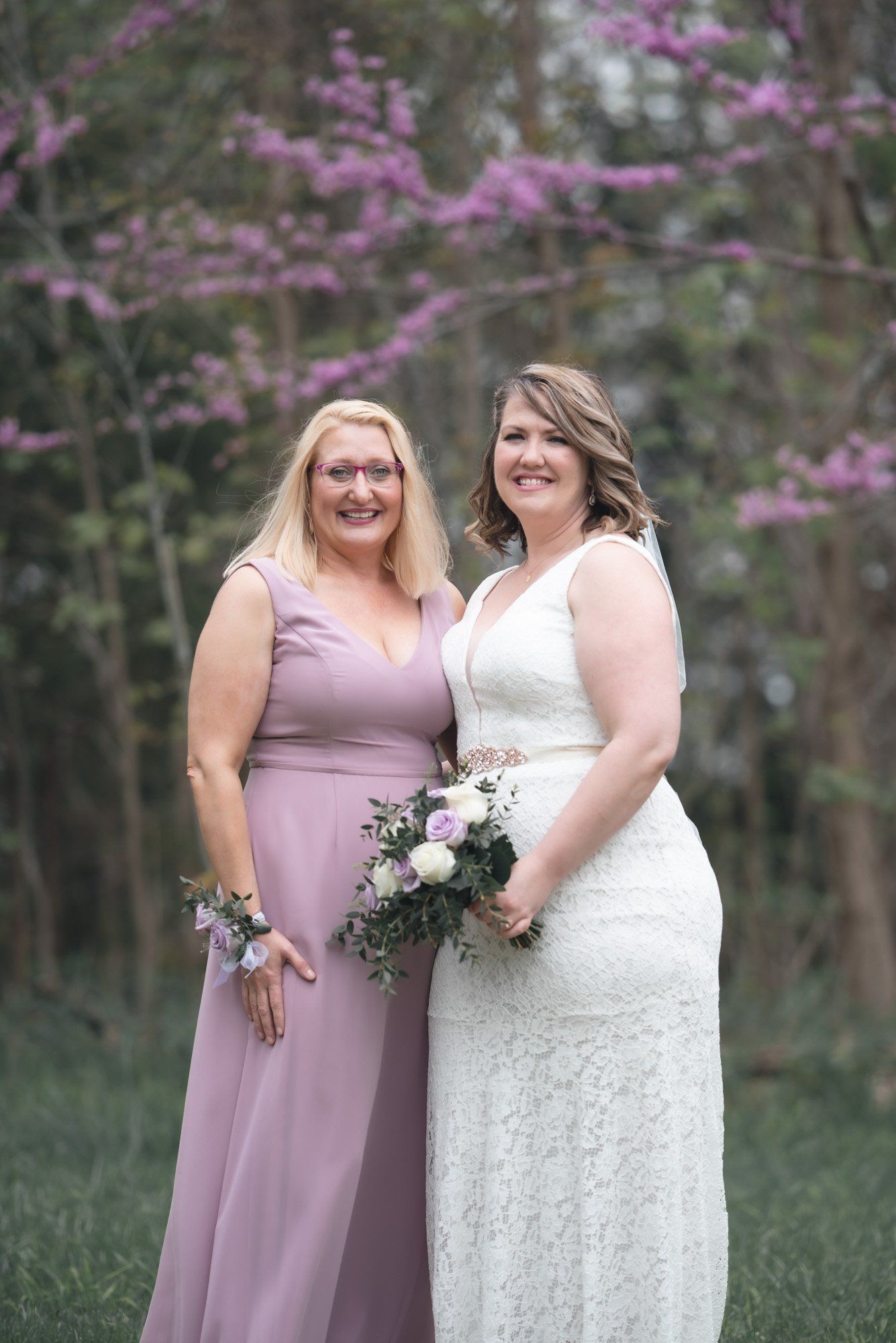 A bride and her bridesmaid are posing for a picture in front of purple flowers.