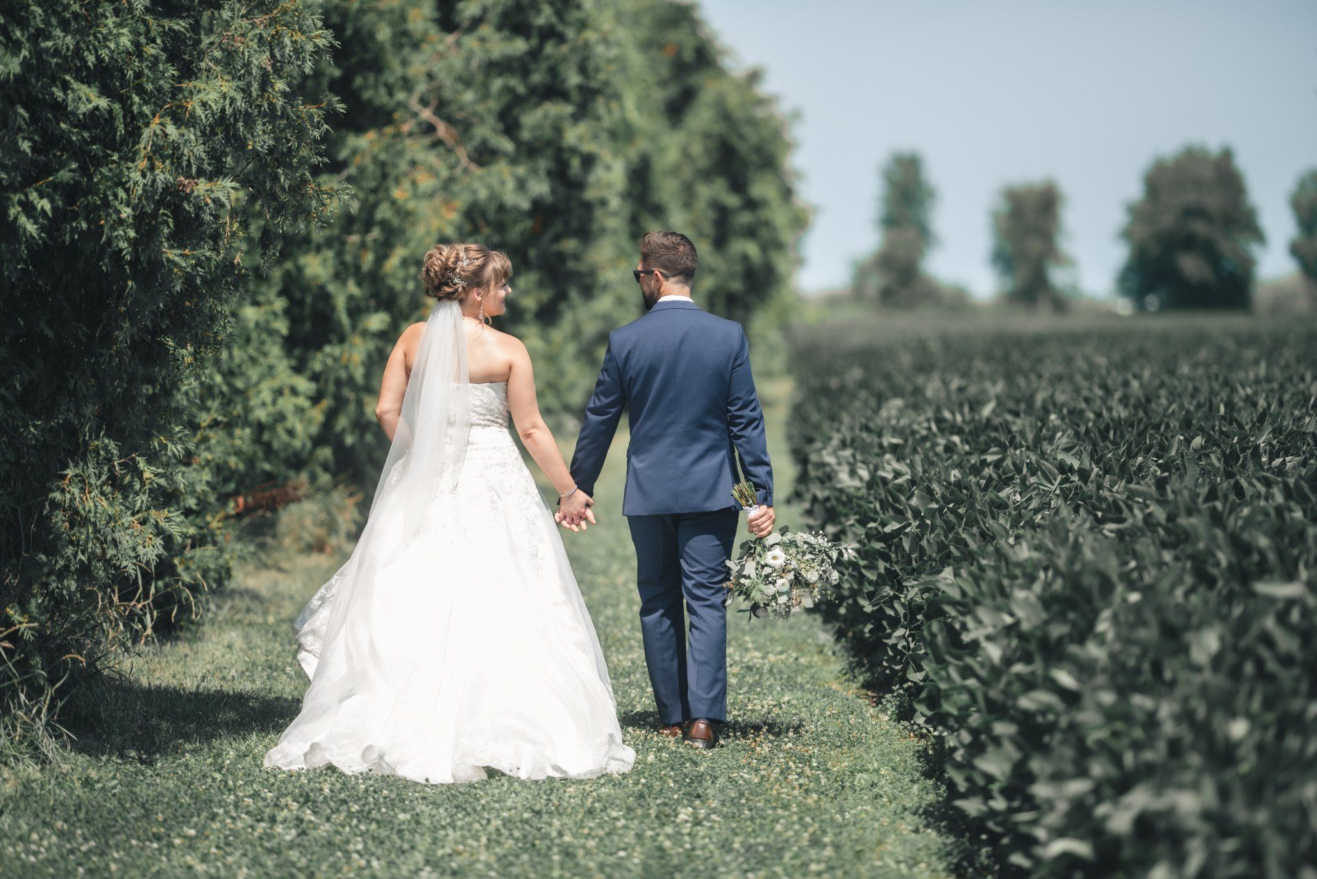 A bride and groom are walking through a field holding hands.
