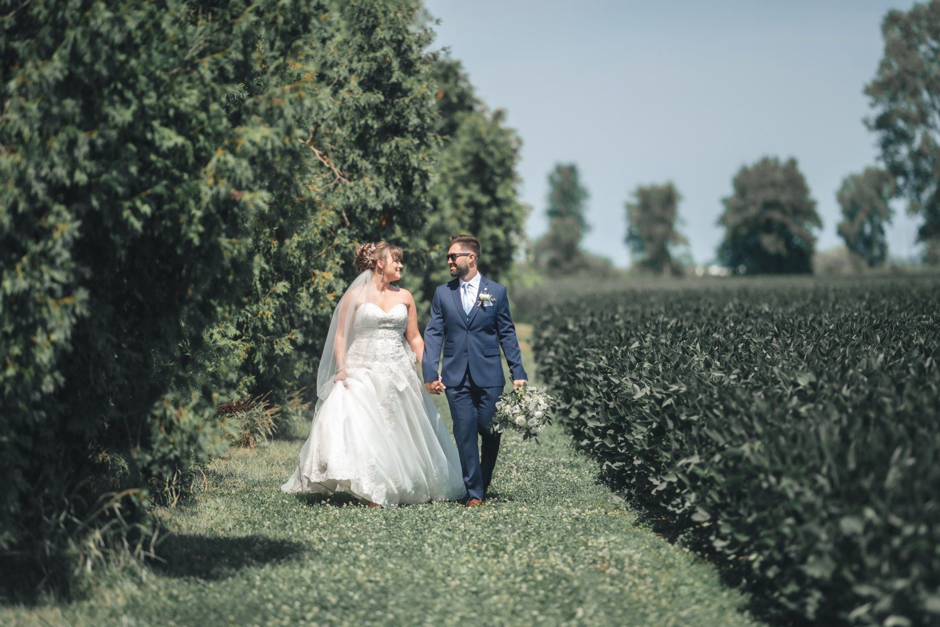 A bride and groom are walking through a field holding hands.