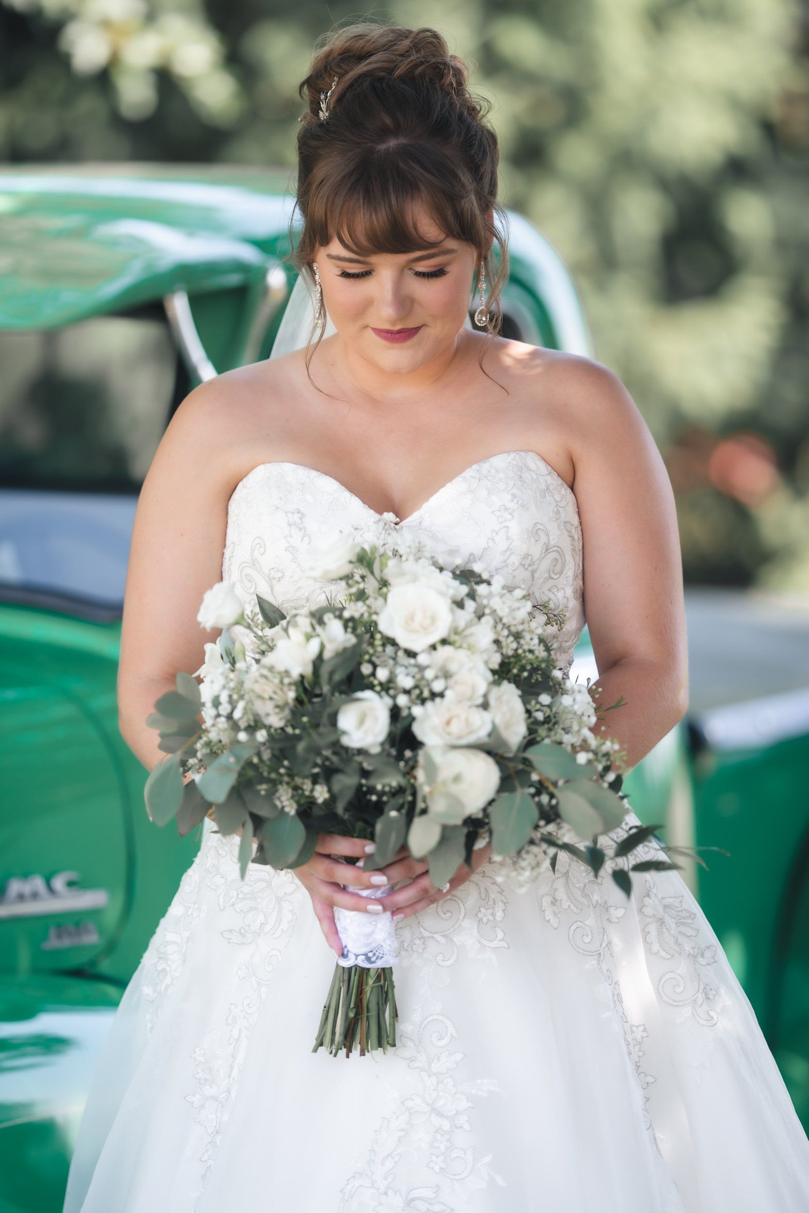 A bride in a white dress is holding a bouquet of white flowers in front of a green car.
