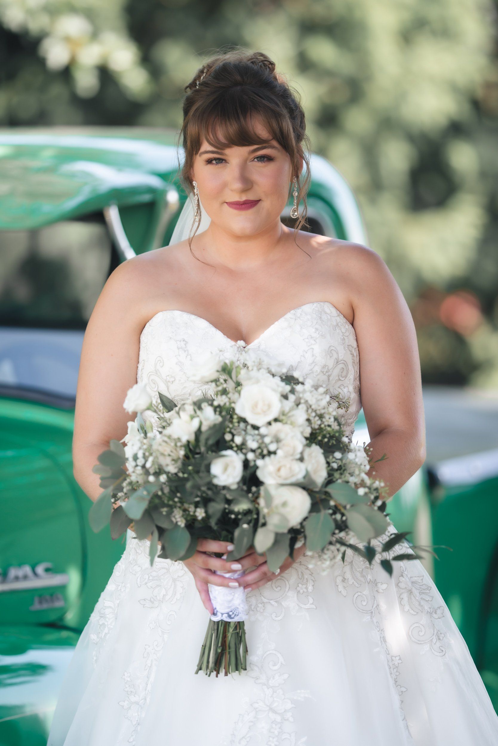 A bride in a white dress is holding a bouquet of white flowers in front of a green car.