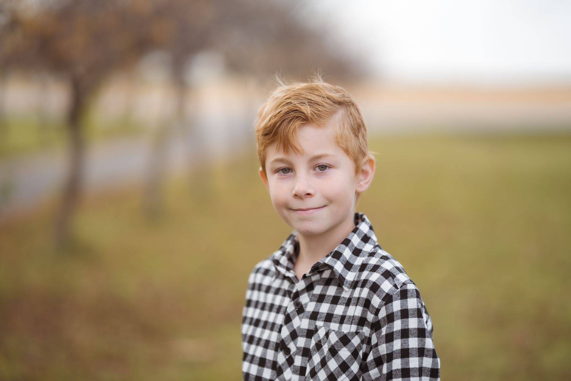 A young boy in a plaid shirt is standing in a field and smiling for the camera.