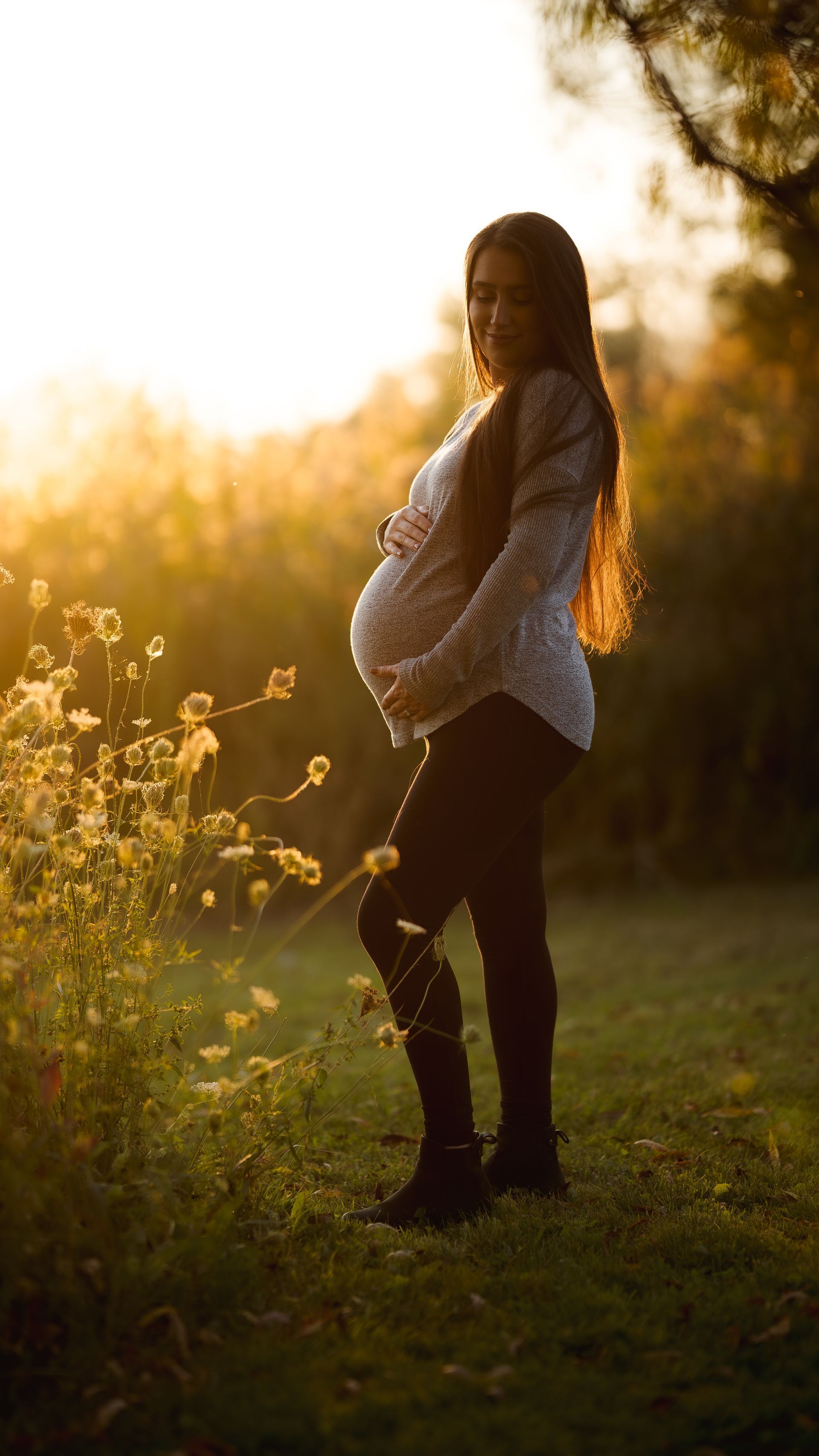 A pregnant woman is standing in a field holding her belly.