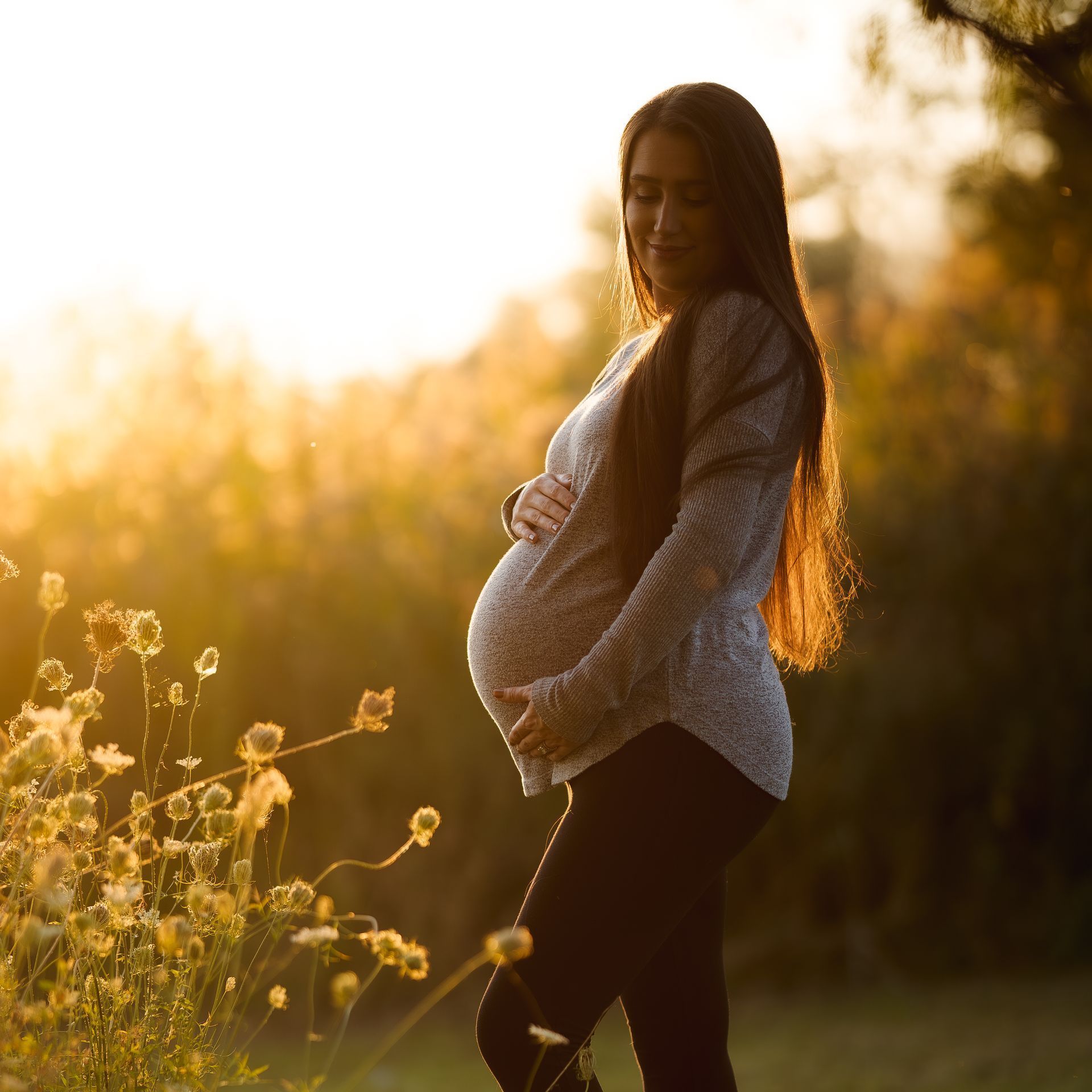 A pregnant woman is standing in a field holding her belly.