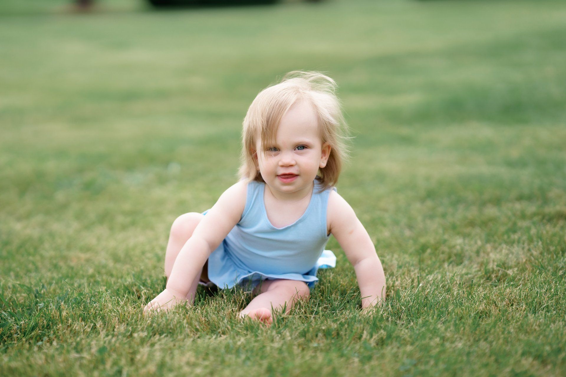 A little girl in a blue dress is sitting on the grass.