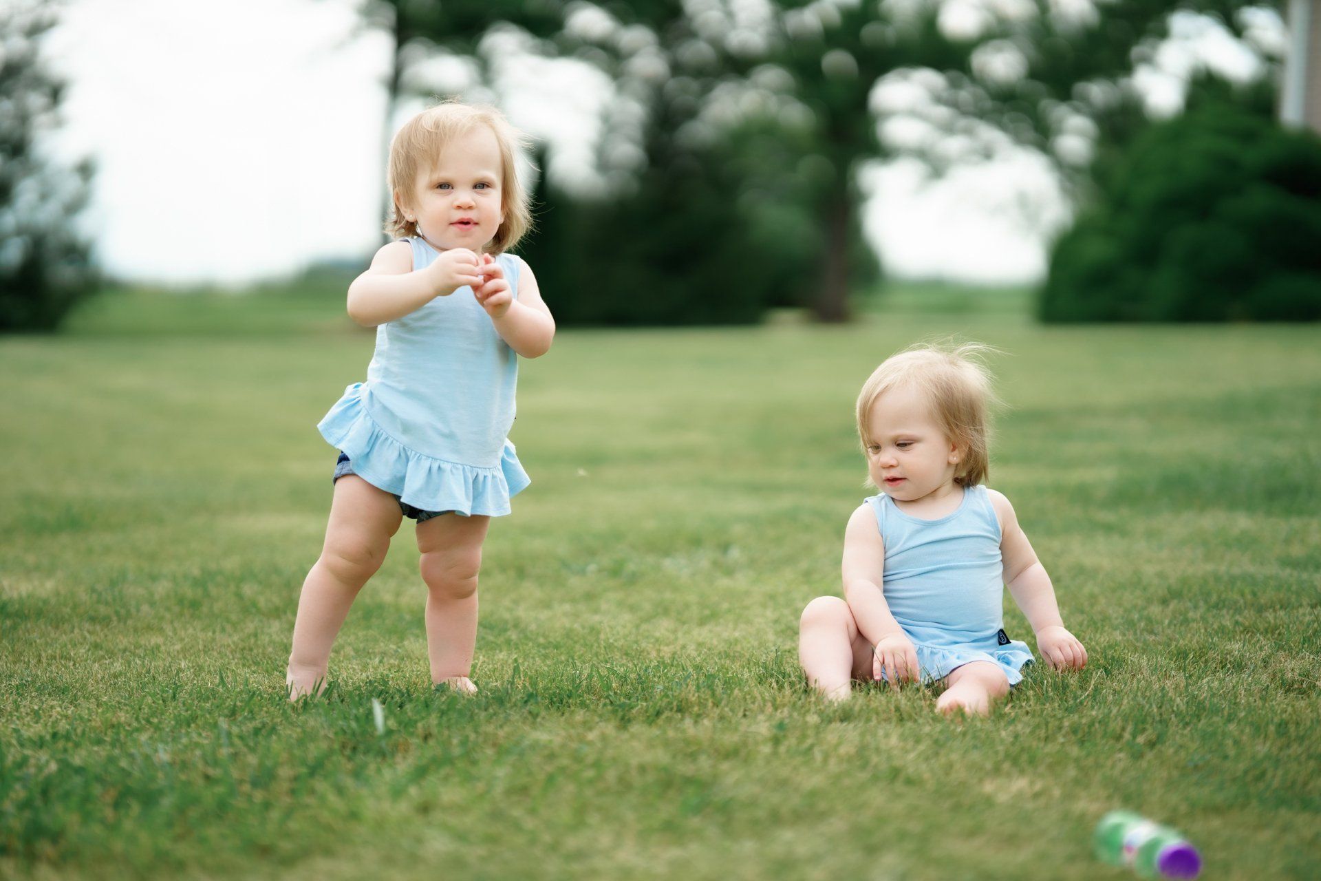 Two little girls are standing and sitting in the grass.
