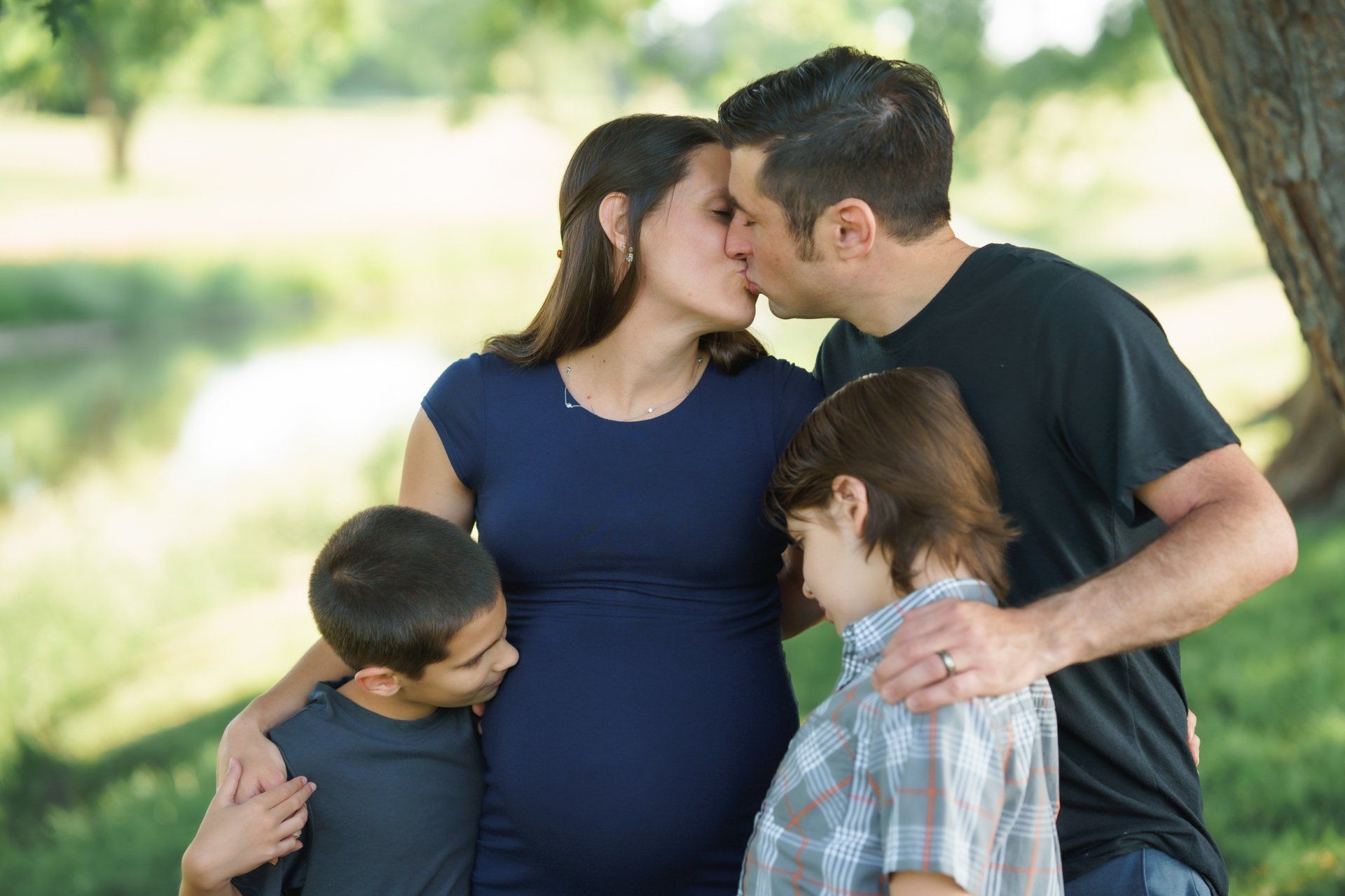 A man is kissing a pregnant woman on the cheek while two boys look on.