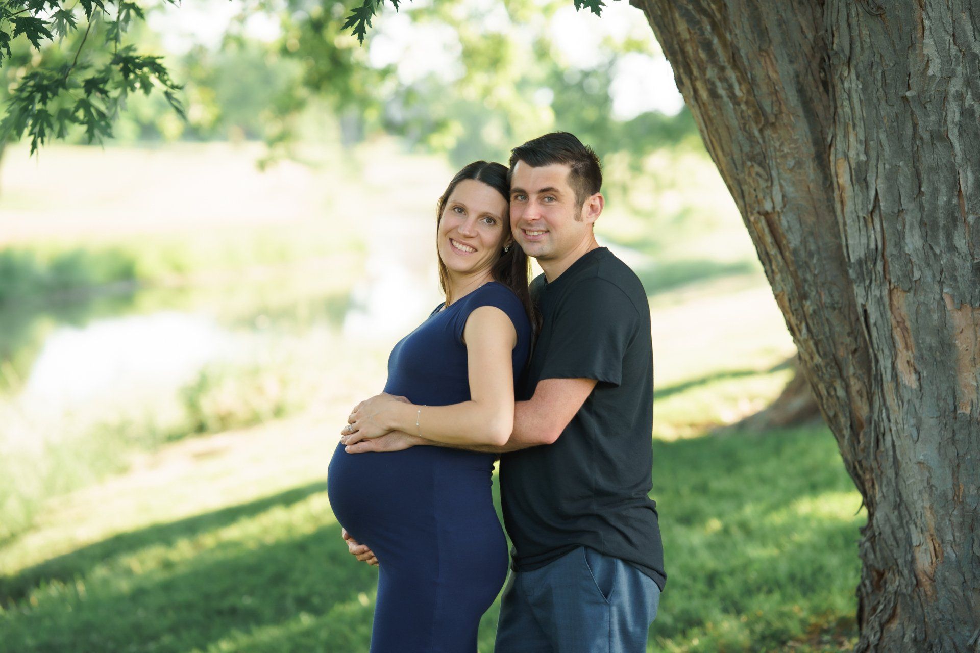 A man and a pregnant woman are posing for a picture under a tree.